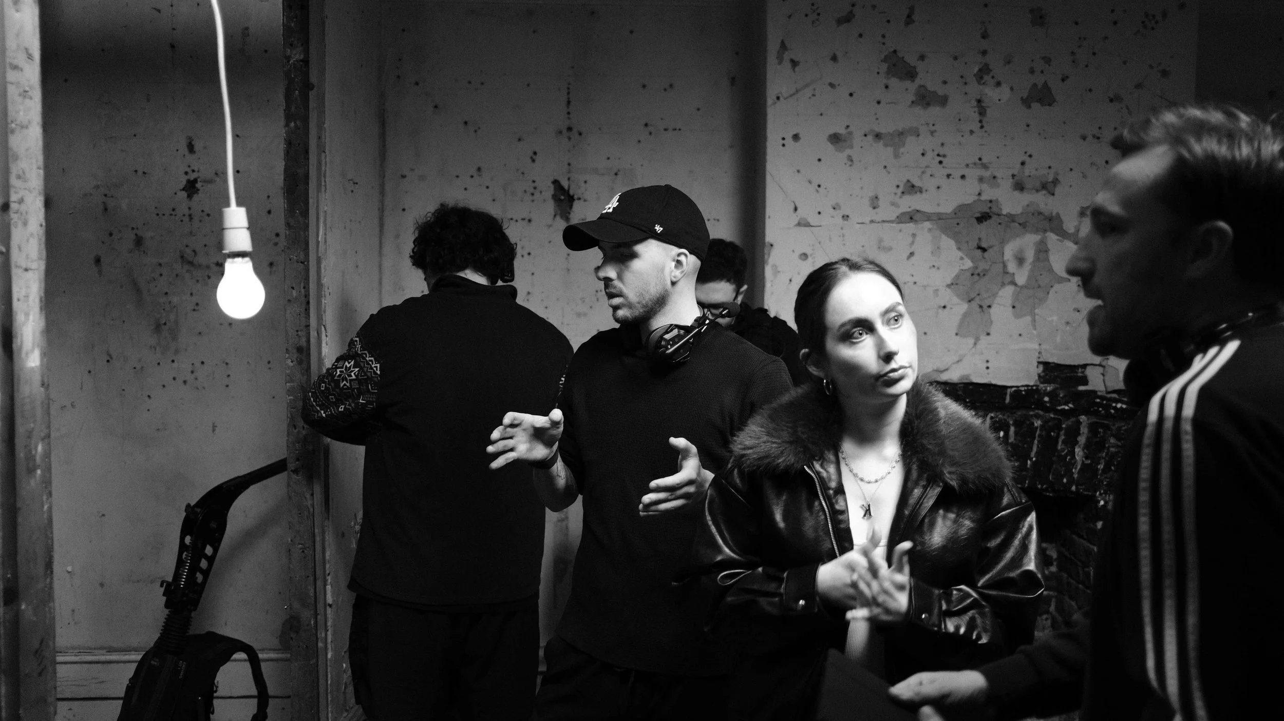A black-and-white photo of five people in a room with a worn, textured wall. One man is wearing a cap and headphones around his neck, gesturing while talking to a woman with dark hair in a leather jacket with a fur collar, who is listening with her h