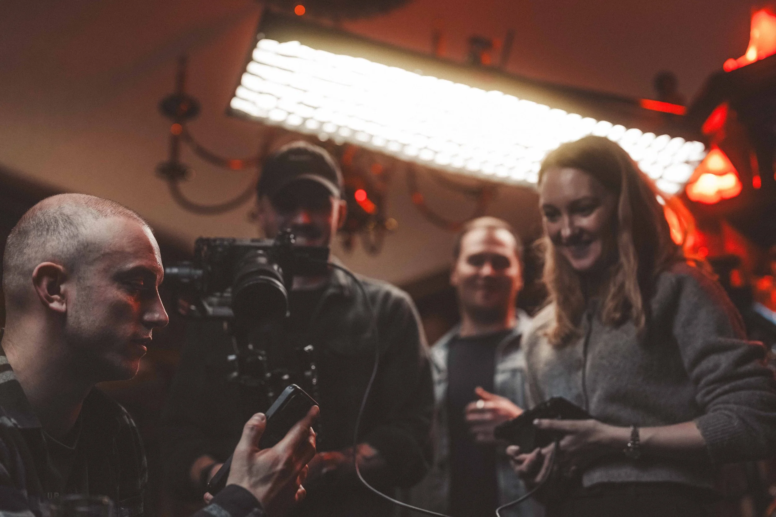 A group of people gathered around a camera, with some looking at their phones, in a dimly lit room with orange and red lighting.