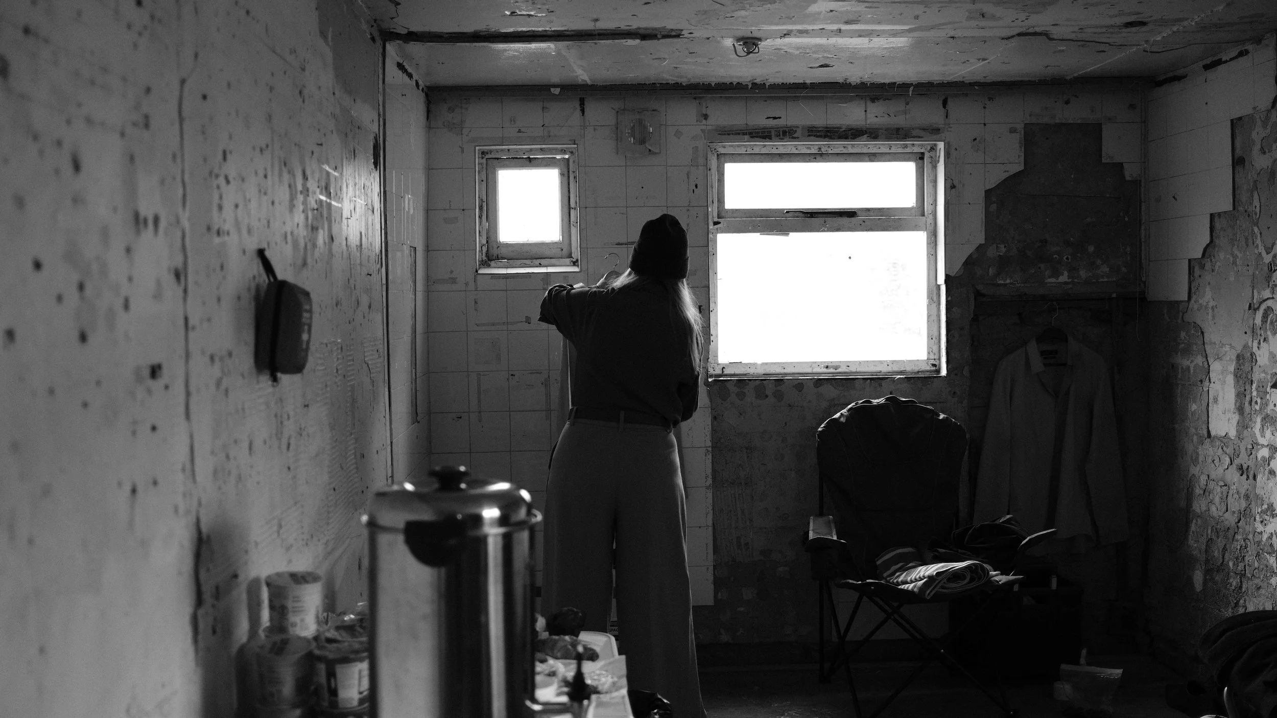 A person standing in a cluttered, dilapidated room with peeling walls and a rough ceiling, looking out a large, bright window.