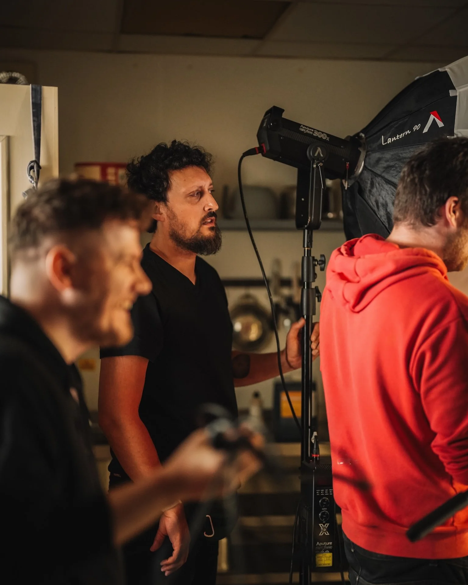 Group of people on a film or photography set, with a man in a black shirt holding a phone, a man in a red hoodie, and a man adjusting lighting equipment, in an indoor setting.