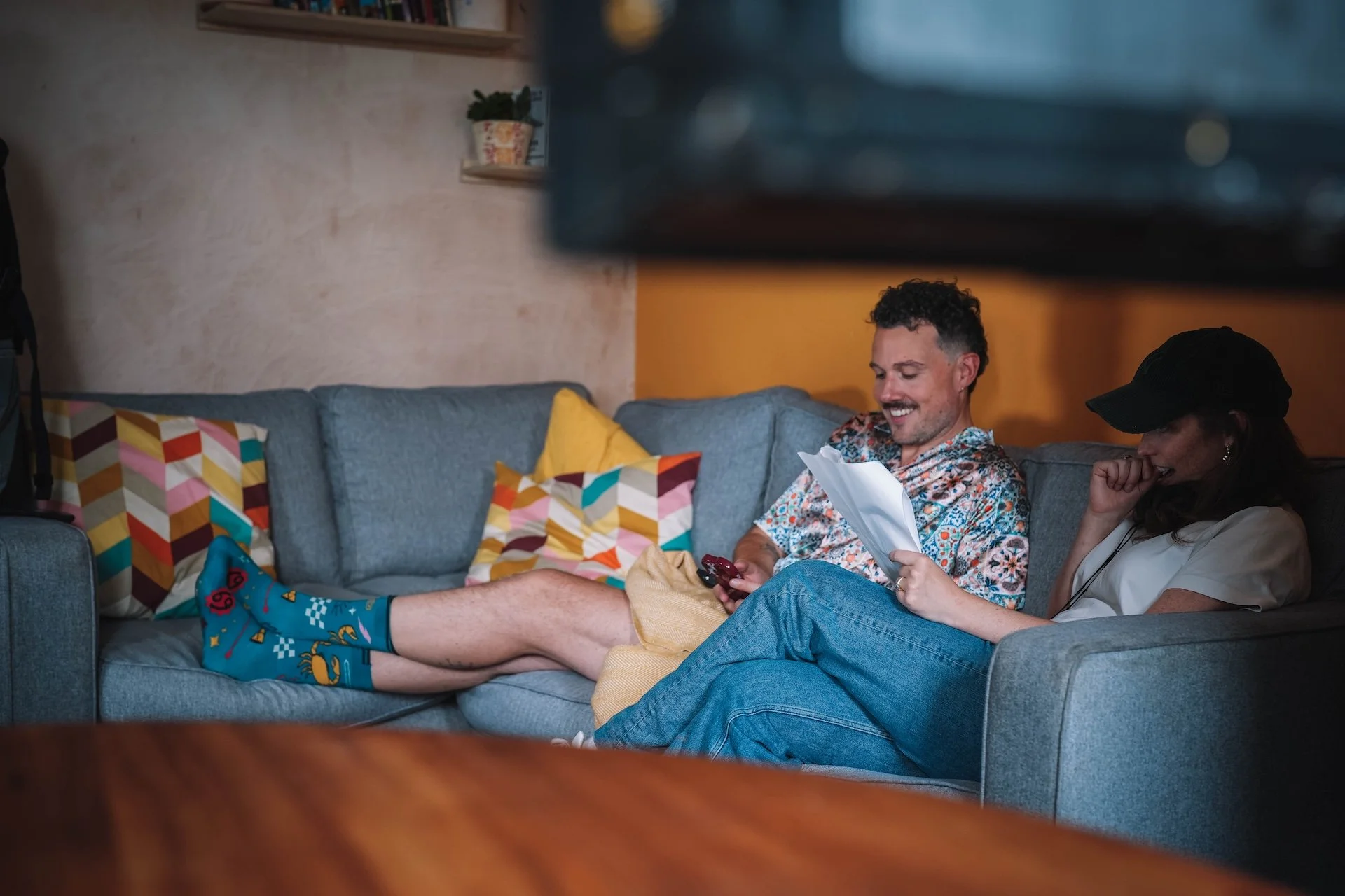 Two people sitting on a gray couch in a living room, one man is lying down with colorful socks and shorts, the woman is wearing a black hat and gray shirt, both are smiling and looking at a phone. The room has colorful pillows and wooden shelves with