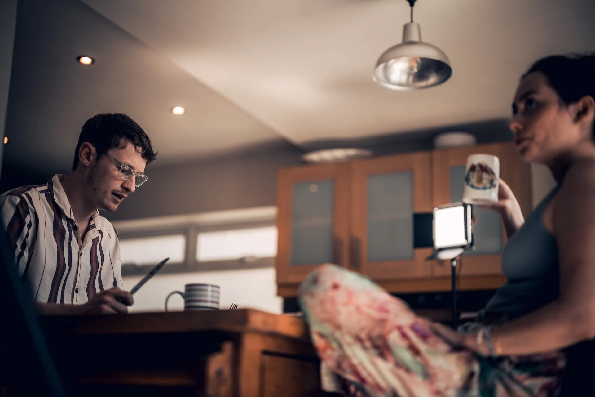 Two people in a kitchen; one man wearing glasses and a striped shirt reading a phone, a woman holding a mug, sitting at a table.