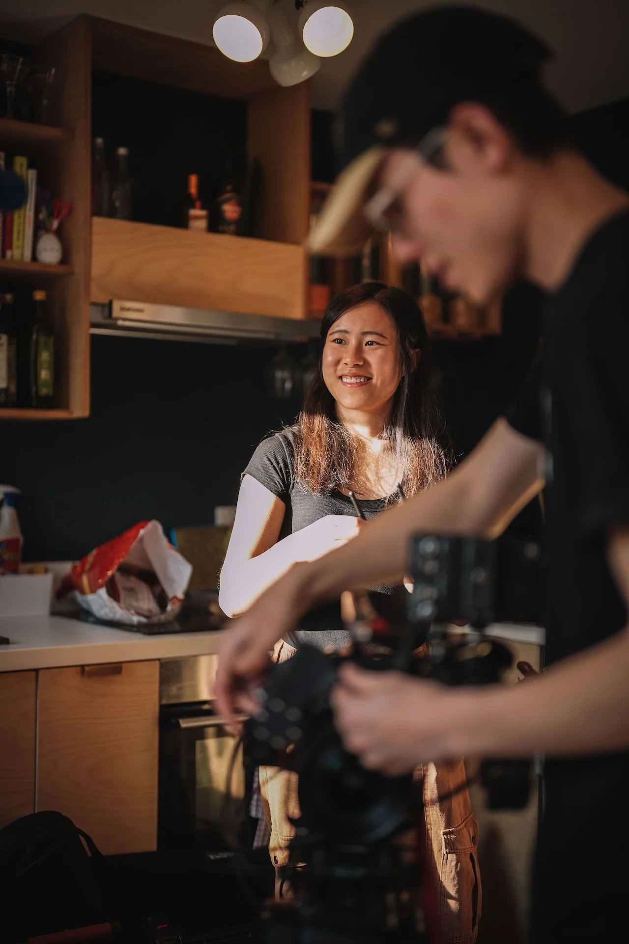 A woman with long brown hair smiling while standing in a kitchen, with a man in glasses and a black cap operating a camera in the foreground.