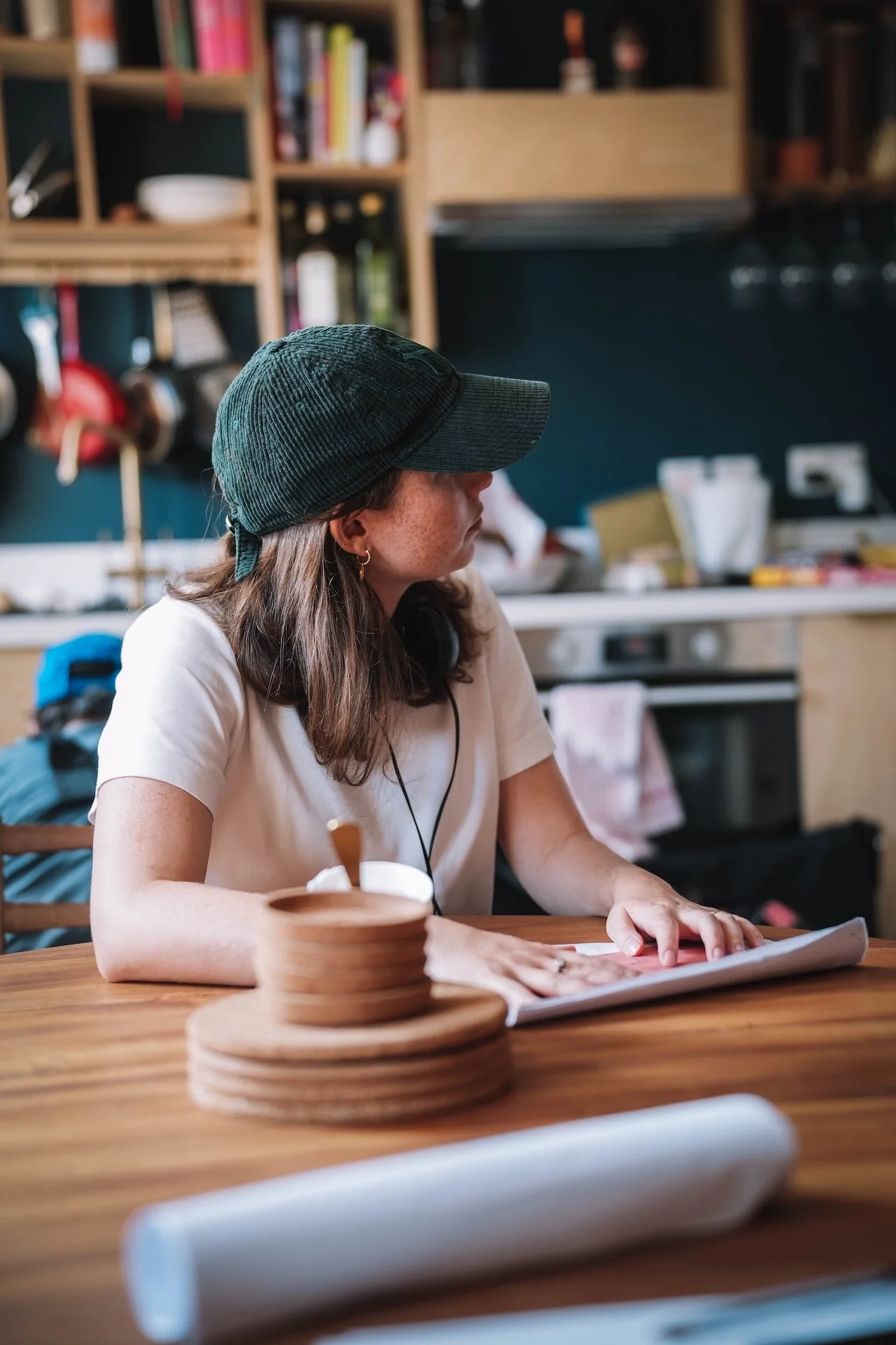A woman with long brown hair wearing a black cap, white shirt, and headphones, sitting at a wooden table reading a magazine in a cozy kitchen with shelves full of books and kitchen items.