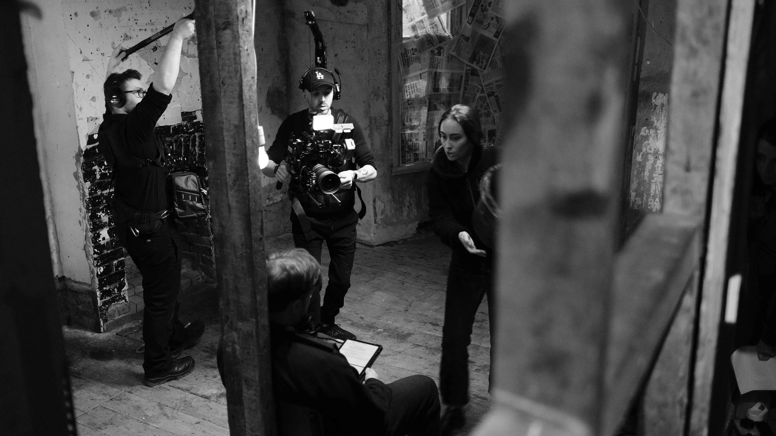 Filming crew setting up a shot in an indoor space with distressed walls and newspaper-covered boards, including a cameraman with headphones, a woman with a clipboard, and a seated man using a tablet.