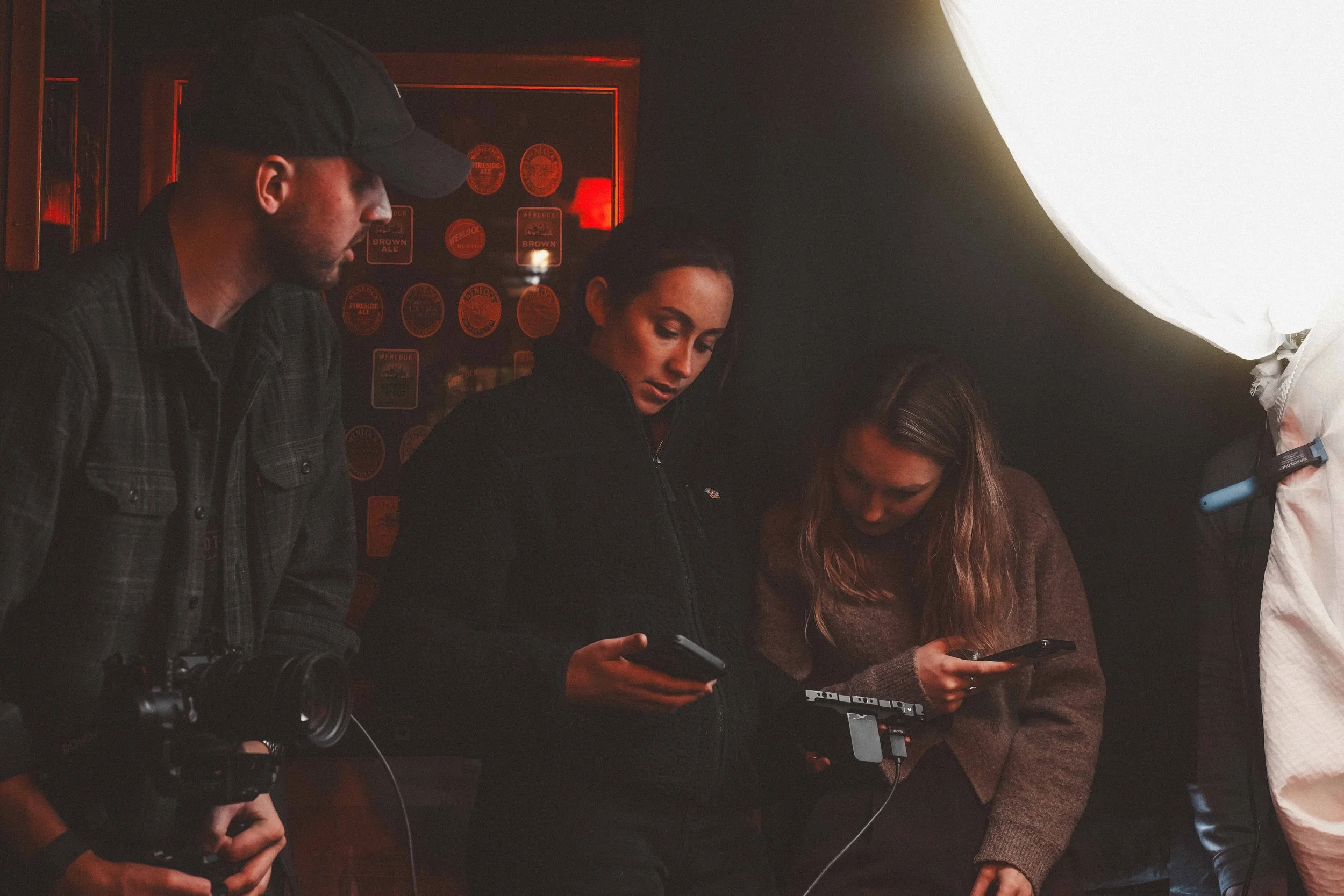 Group of four people standing together indoors, looking at their smartphones. One person holds a camera, others are focused on their devices. Background has red and black decor and illuminated signs.