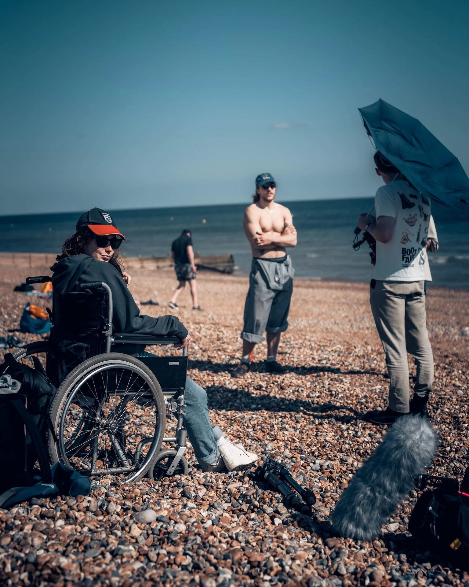 People on a pebbled beach, including a woman in a wheelchair wearing sunglasses and a cap, a shirtless man with crossed arms, and another person holding an umbrella and filming. In the background, the ocean extends to the horizon with a clear sky.