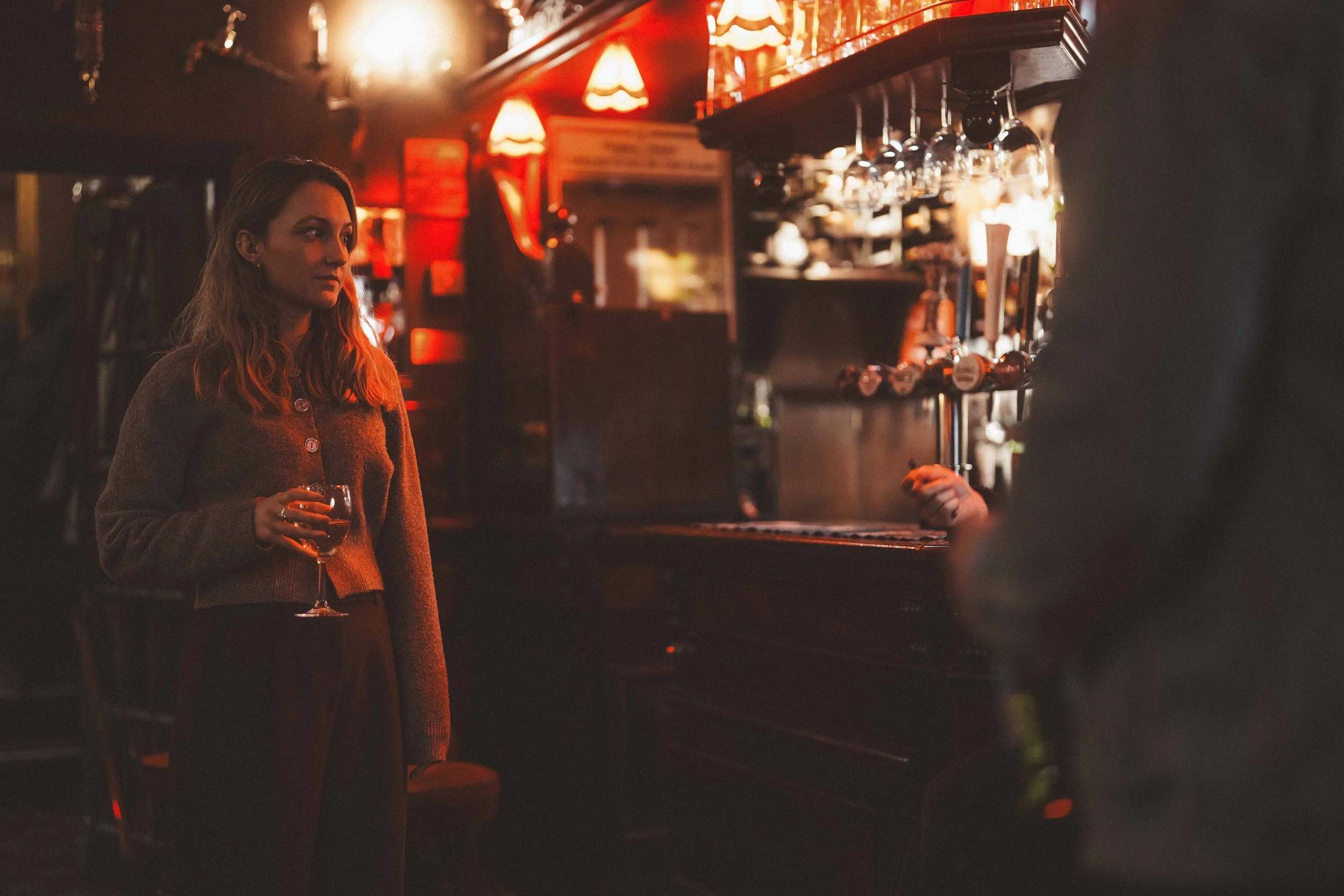 A woman with wavy hair holding a glass of wine, standing in a dimly lit bar or pub with warm red lighting and hanging glasses above the bar counter.