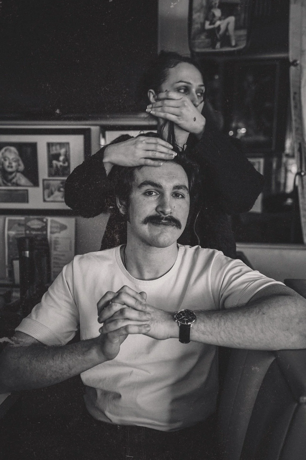 A man with a mustache and wristwatch sitting at a table, with a woman standing behind him, covering part of her face with her hand, in a room decorated with framed pictures on the wall.
