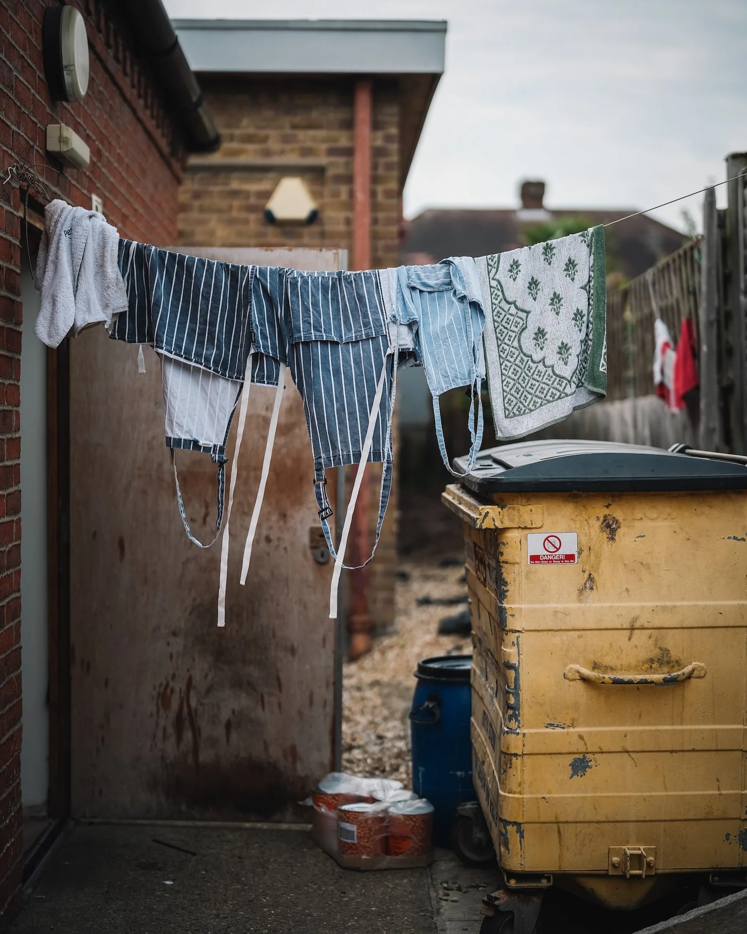 Clothes hanging on a clothesline and trash bin in alleyway of a residential backyard.