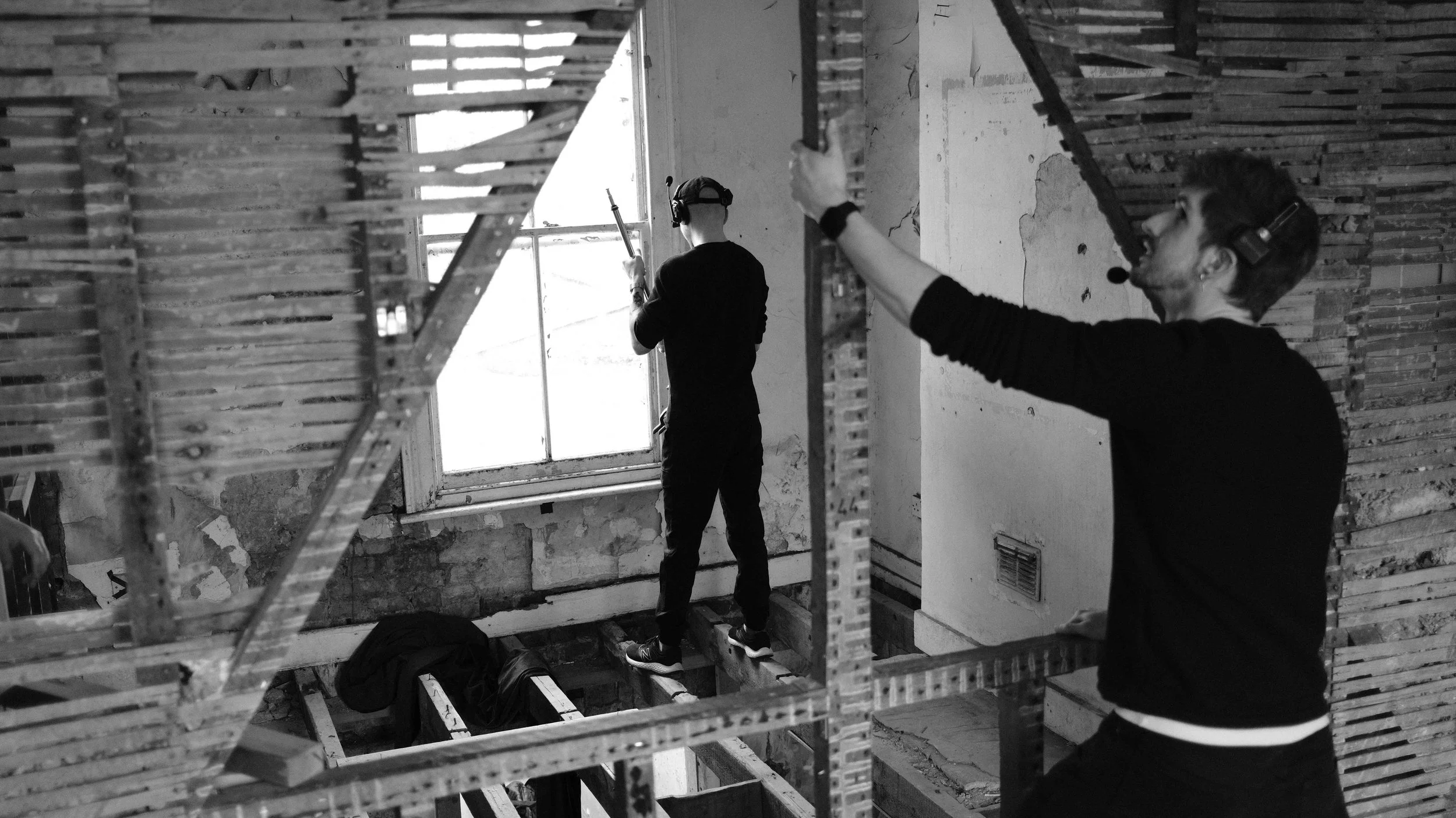 Two workers on a construction site, with one standing by a window using a tool and another directing or signaling with their hand, surrounded by scaffolding and exposed walls.