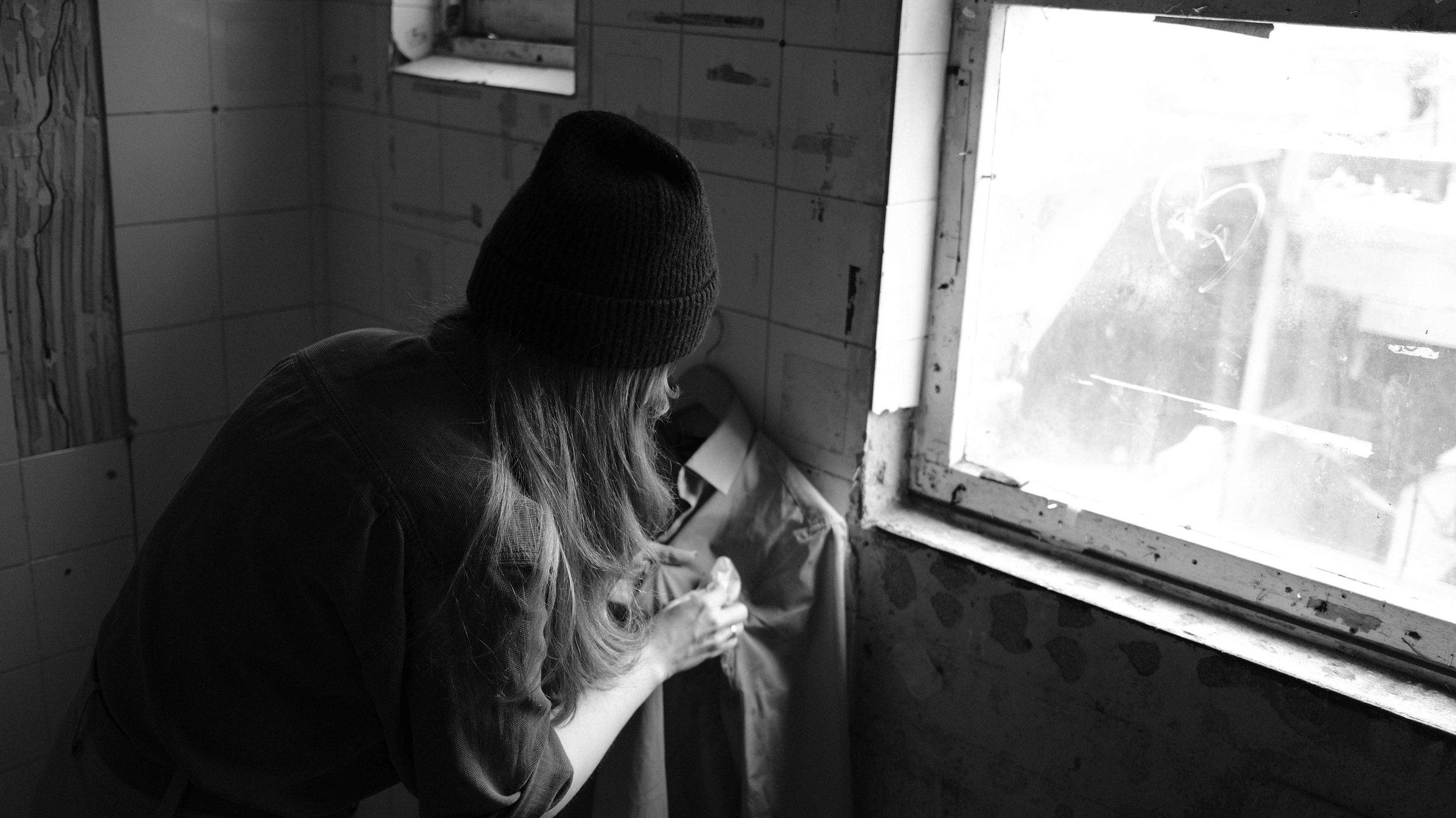 A woman wearing a knit hat and a jacket is looking down, interacting with a bag or package near a window in a room with tiled and rustic wall surfaces, in black and white.