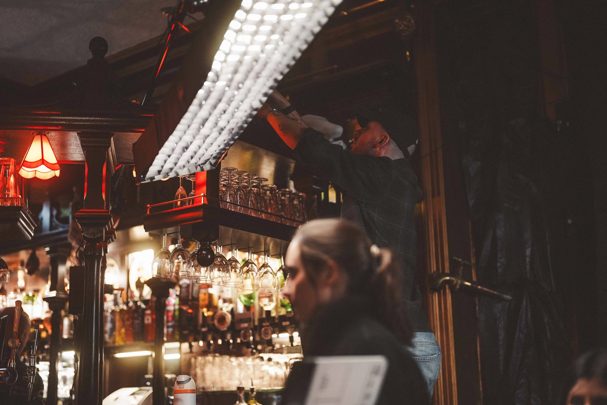A bartender pouring a drink behind a bar counter in a dimly lit bar with glassware and bottles on shelves, warm lighting, warm wooden interior, and patrons in the foreground.