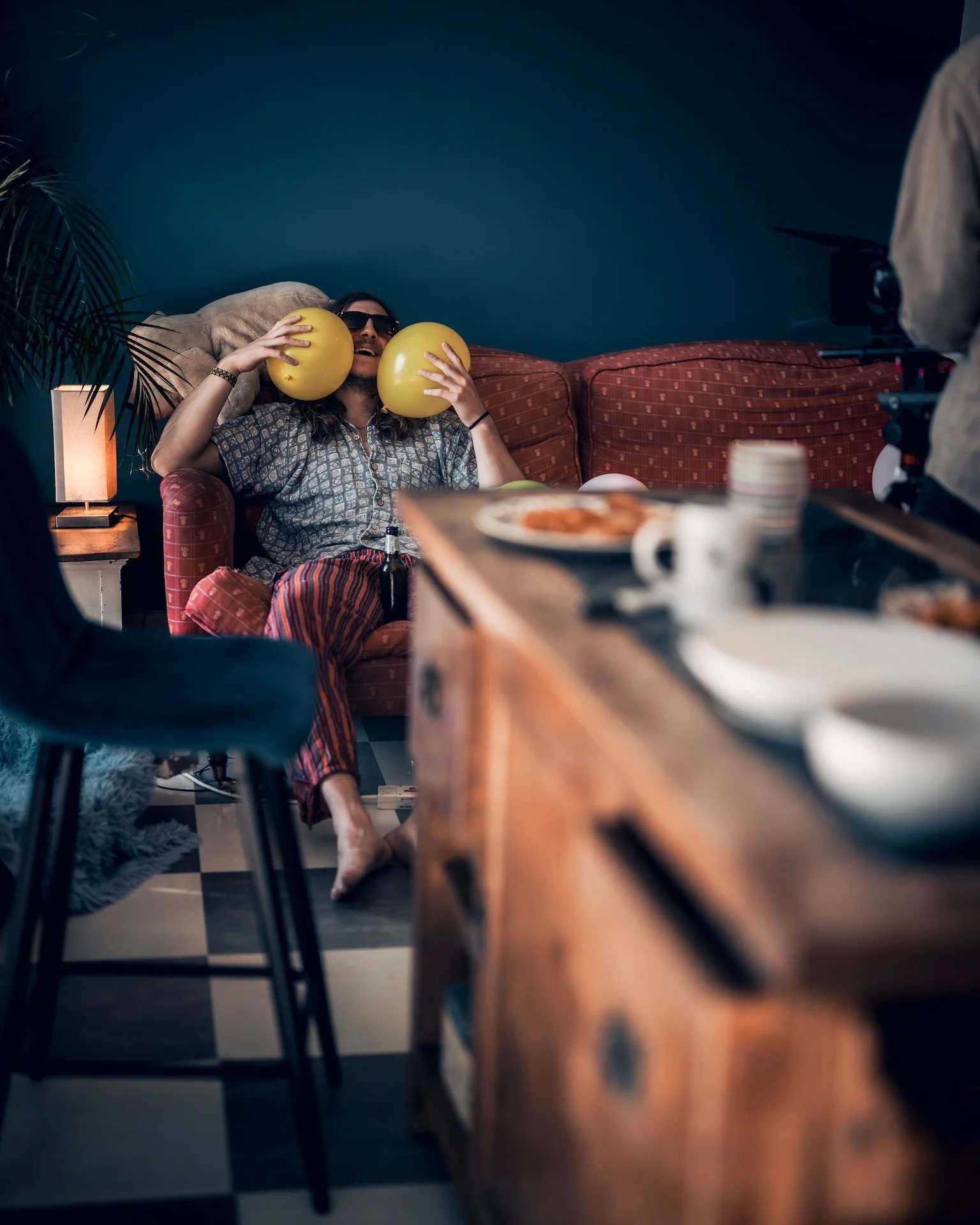 Man sitting on sofa with sunglasses, holding yellow balloons near his face, in a dimly lit room with a teal wall, table with food and drinks in the foreground, and a camera operator filming him.