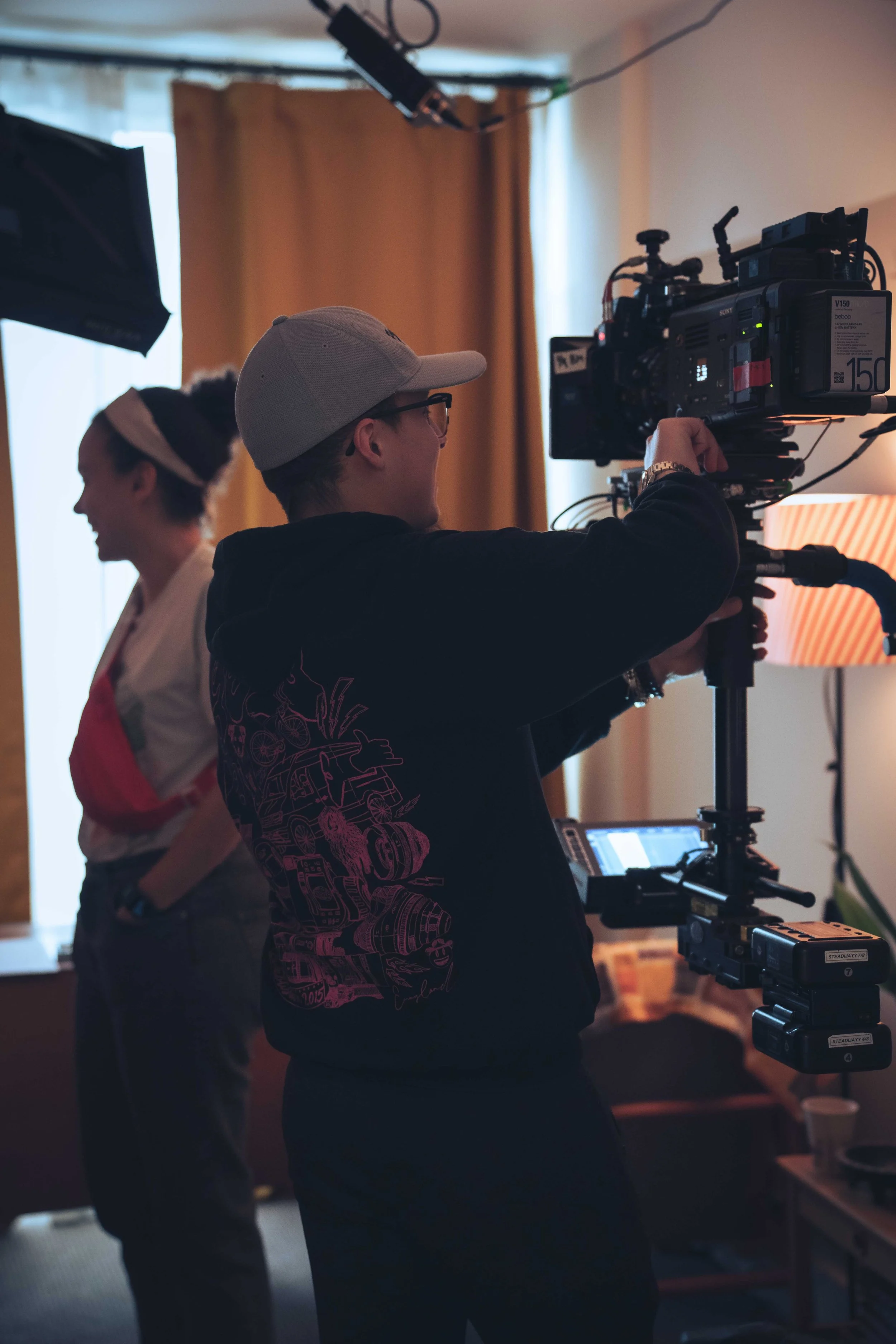 A person operating a professional film camera indoors with another person in the background, both women, one wearing a white kimono-style outfit and the other in casual clothing. The scene appears to be part of a film or video production.