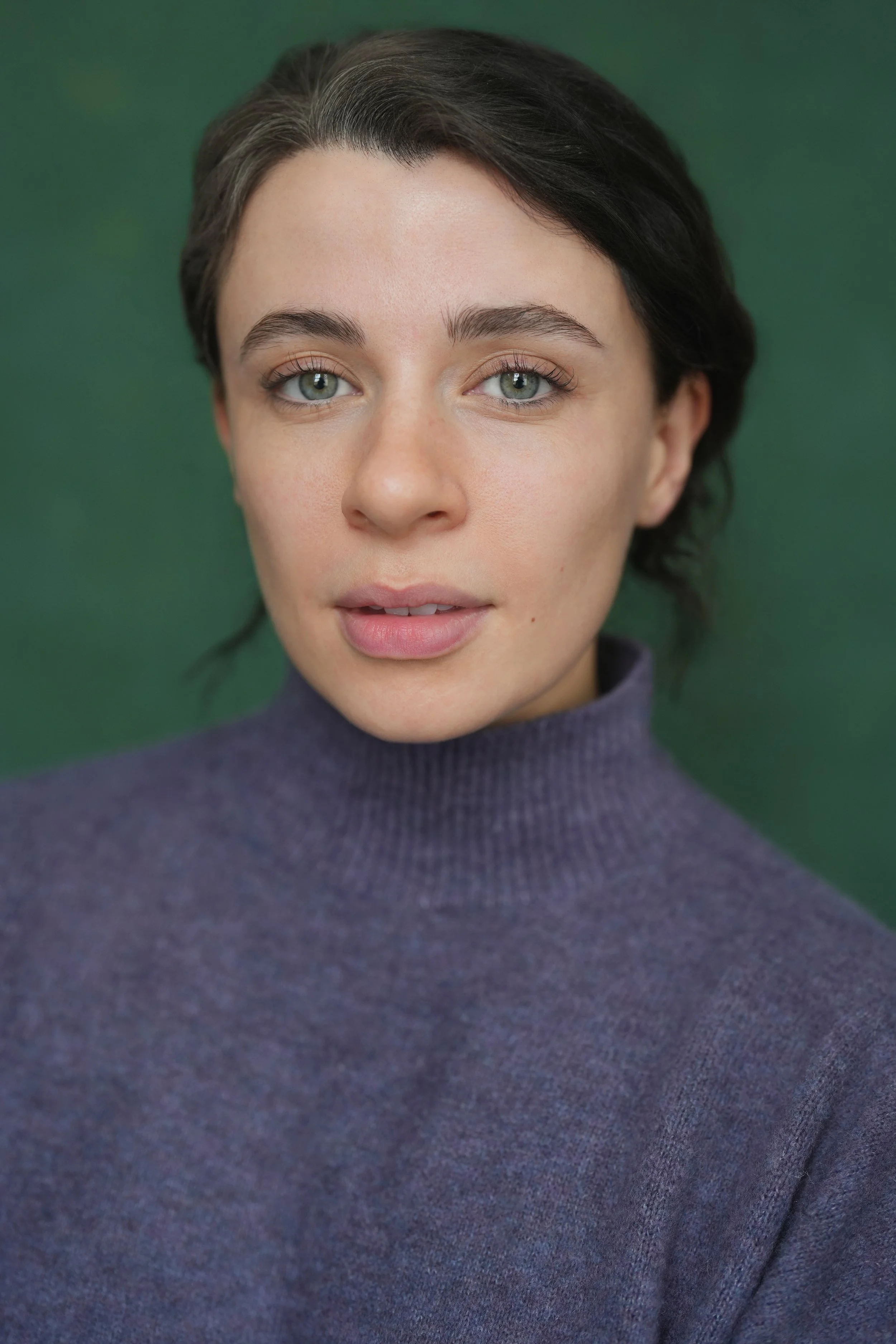 Close-up portrait of a woman with short dark hair, blue eyes, wearing a purple turtleneck sweater, against a green background.