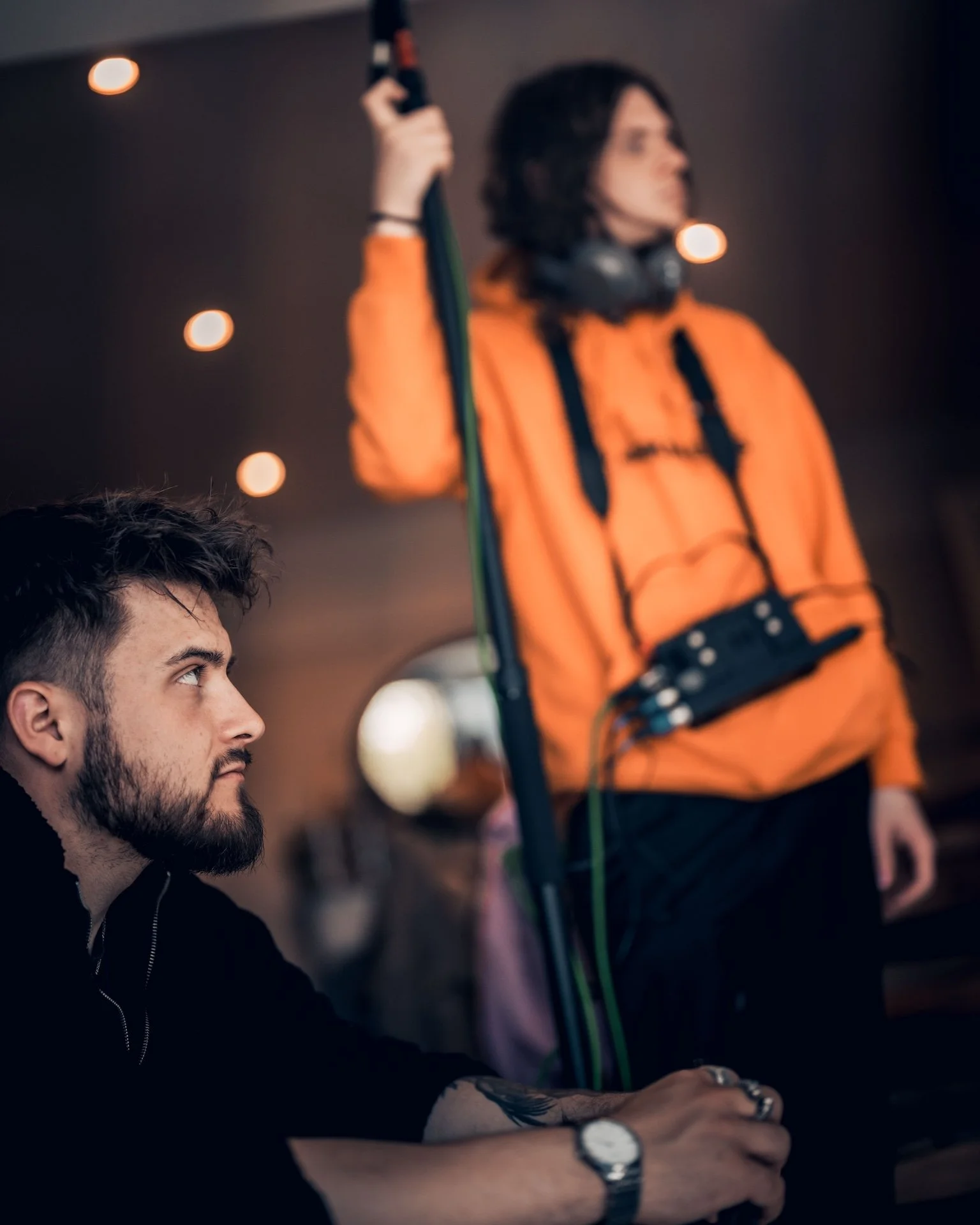 A man with a beard and tattoos on his arm, wearing a black hoodie, sitting at a table, and looking intently at something off-camera. In the background, a woman with dark hair, wearing an orange jacket and headphones around her neck, is holding a micr