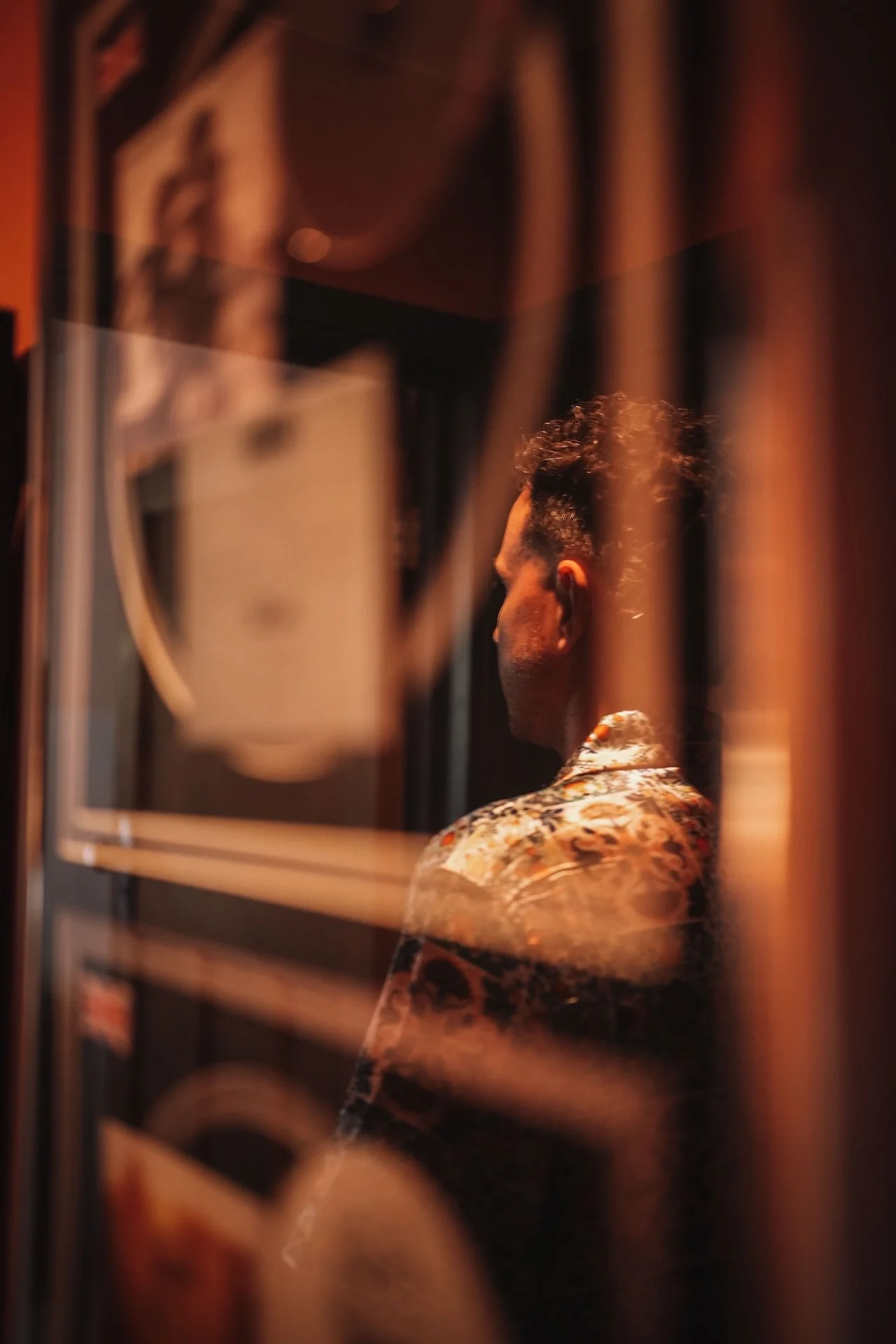 A person with curly hair wearing a patterned shirt, viewed from behind, seen through a blurred metal rack or grid.
