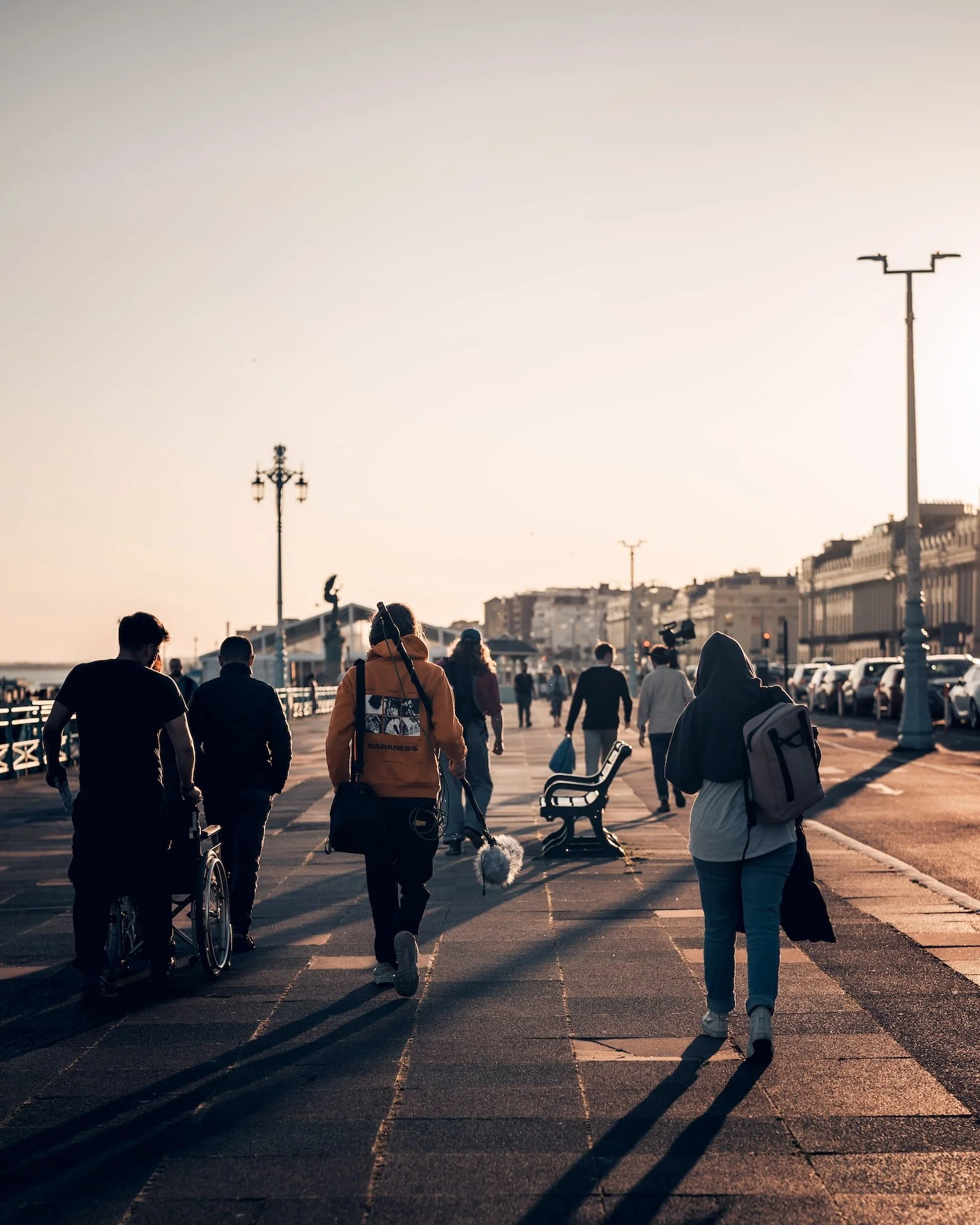 People walking along a city pier during sunset, with the sea to the left and buildings in the background.