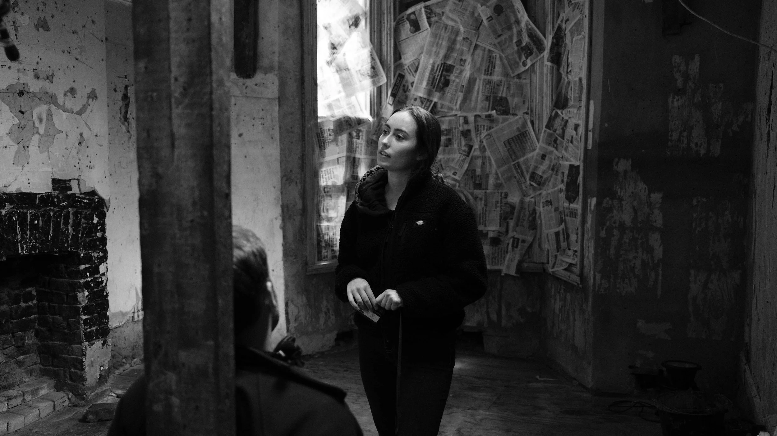 A woman standing in a dilapidated room, talking to a seated person whose face is not visible. The room has peeling walls and a window covered with newspapers.