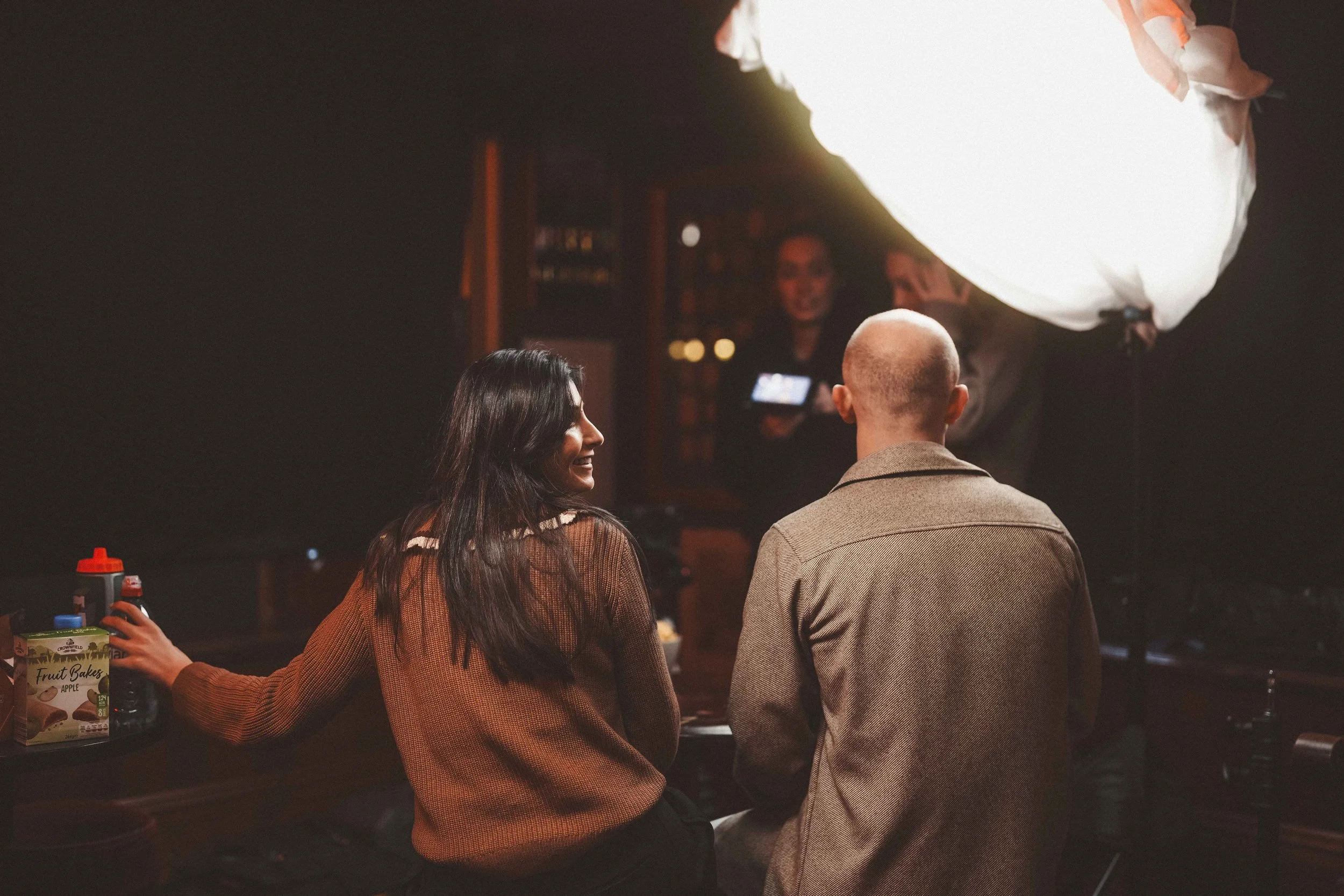 A woman and a man sit together at a social event, smiling and talking. In the background, a person holds a camera, and a large illuminated paper lantern hangs overhead. A table next to them has bottles and juice cartons.