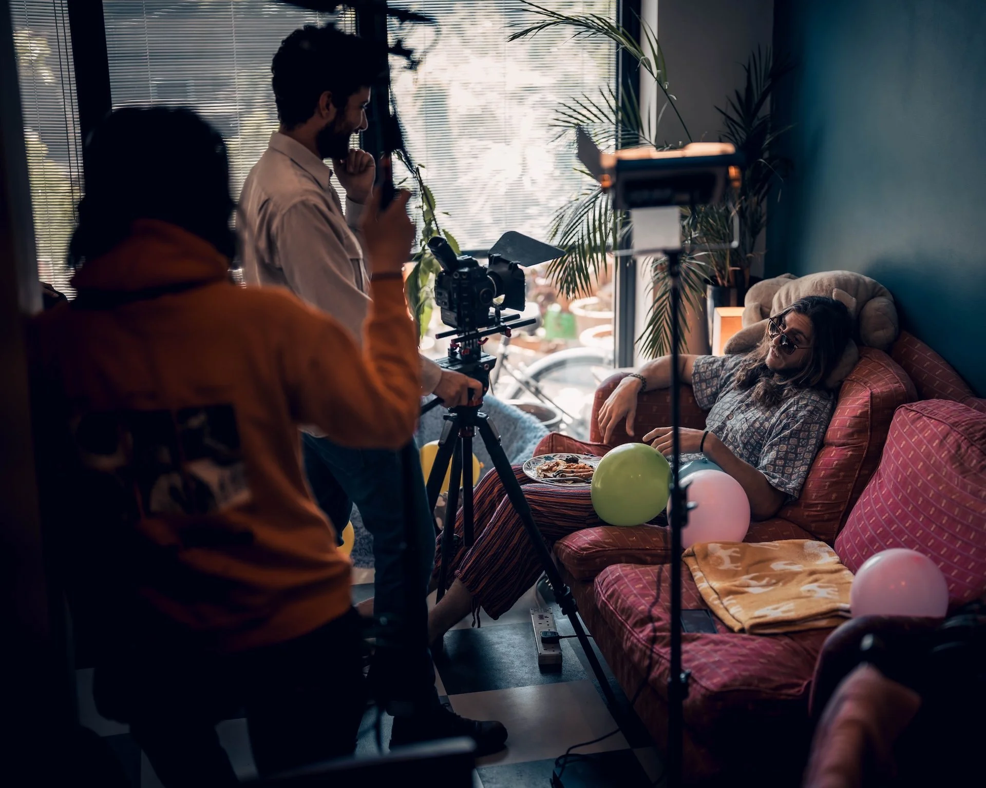 A woman with sunglasses relaxing on a sofa during a video shoot, with balloons around her. Two men are filming her with camera equipment in front of a window with blinds, in a room decorated with large indoor plants.