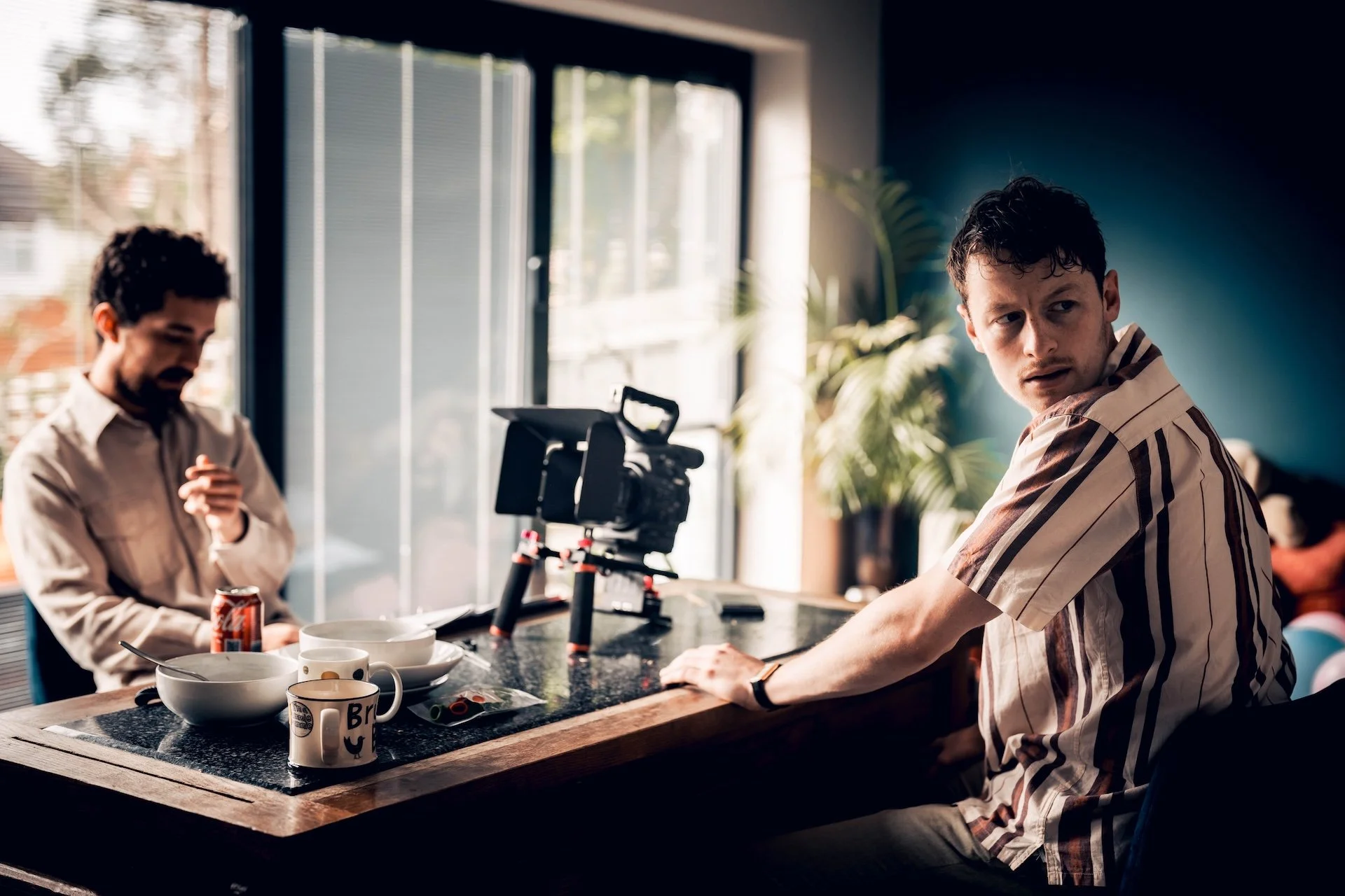 Two men working in a studio with a camera, bowls, and mugs on the counter, one man working on a project and another looking to the side.