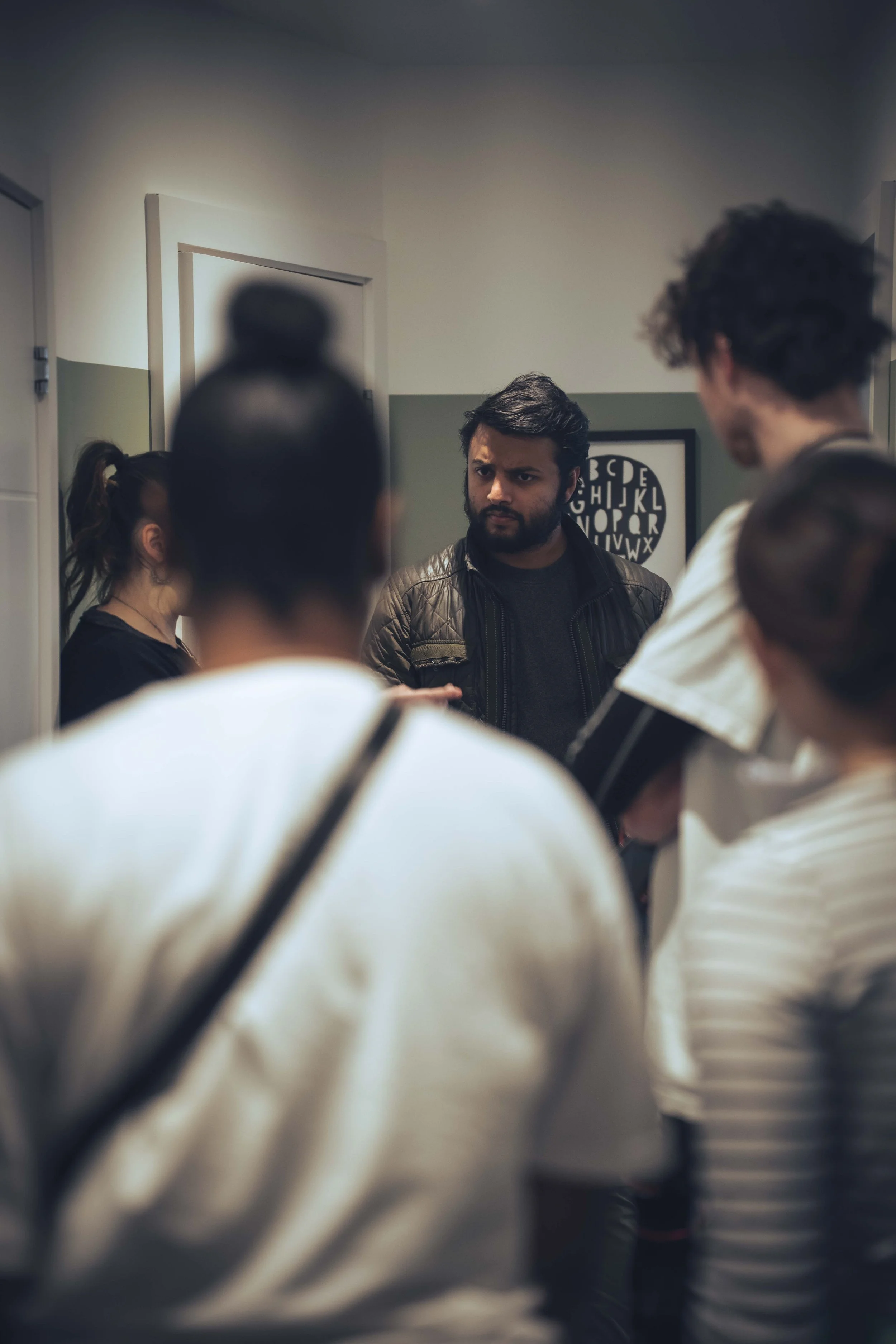 A man with dark hair and a beard, wearing a black jacket, looks serious as he interacts with a group in a room with gray and white walls. Several people are partially visible around him, some with blurred faces. There is a framed poster of the alphab