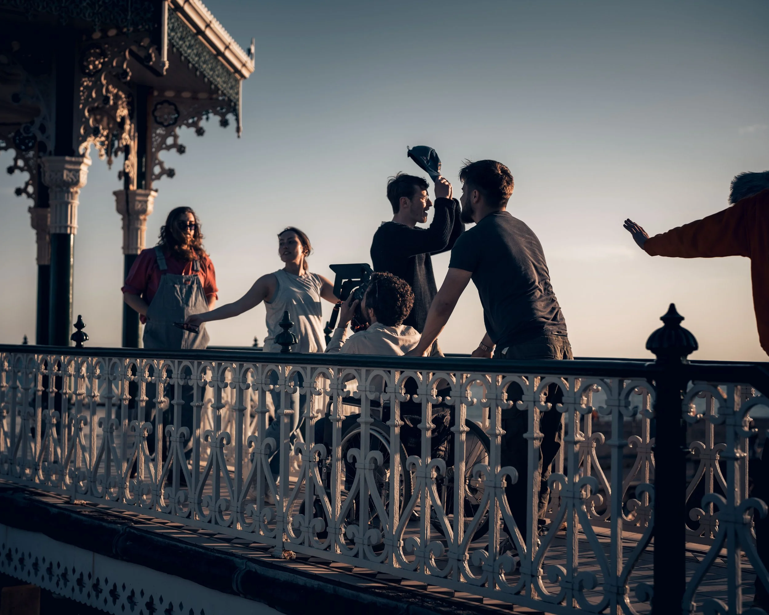 People standing and talking on a rooftop deck with ornate white railing, an ornate gazebo structure in the background, and a clear sky.