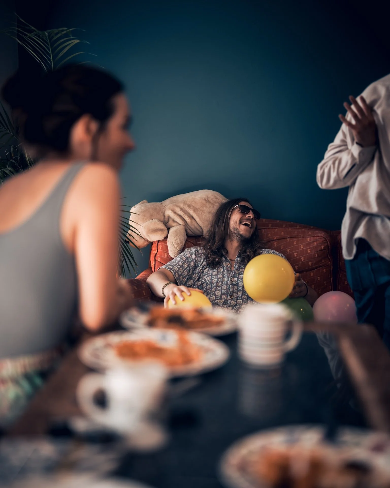 People celebrating at a party with balloons and food, one person sitting on a sofa wearing sunglasses and laughing, others standing around.