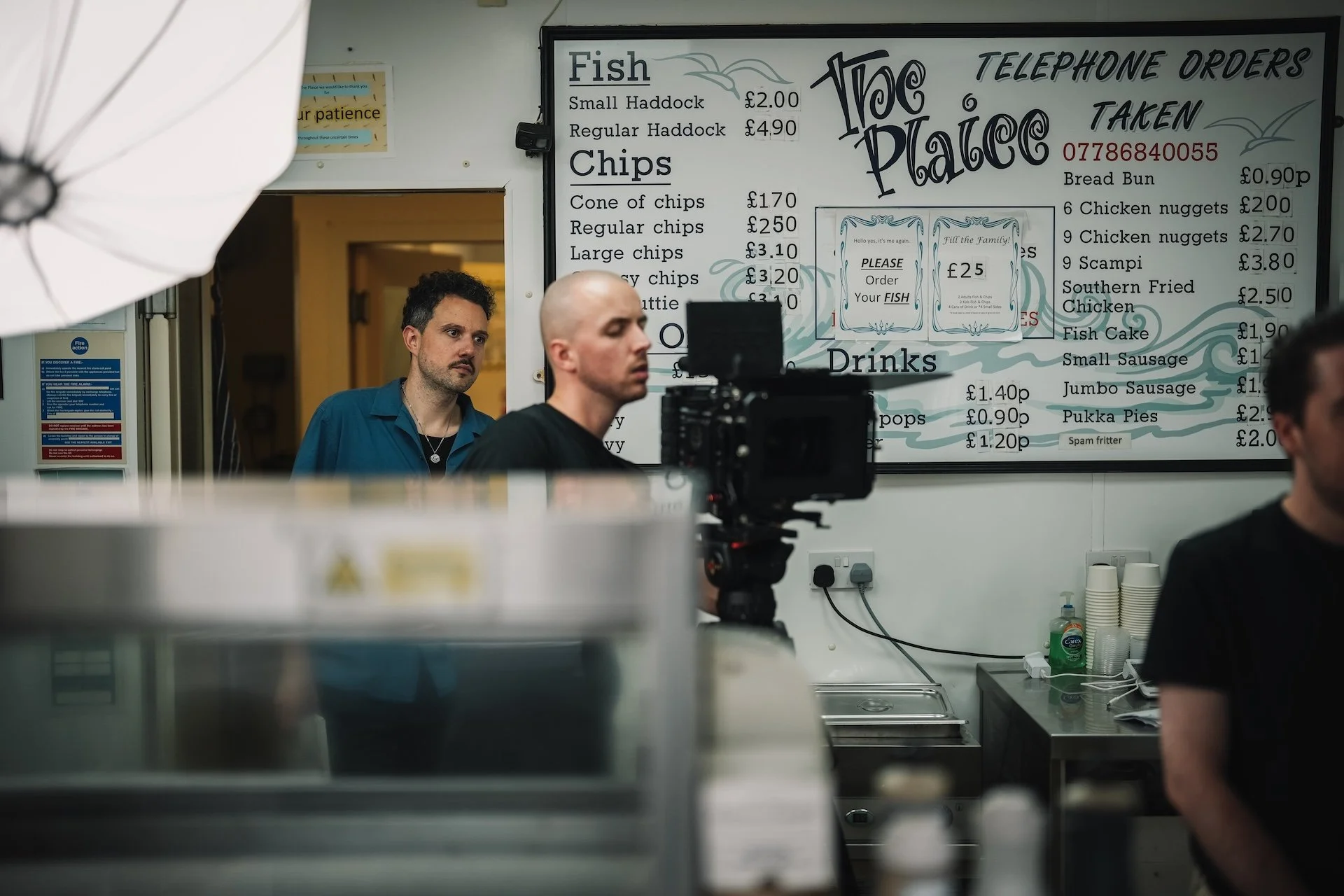 Inside a fish and chips shop with menu board and staff. The menu board lists different sizes of haddock and chips, with prices. There are other food items and prices listed, as well as a sign for telephone orders taken. Staff are visible behind the c