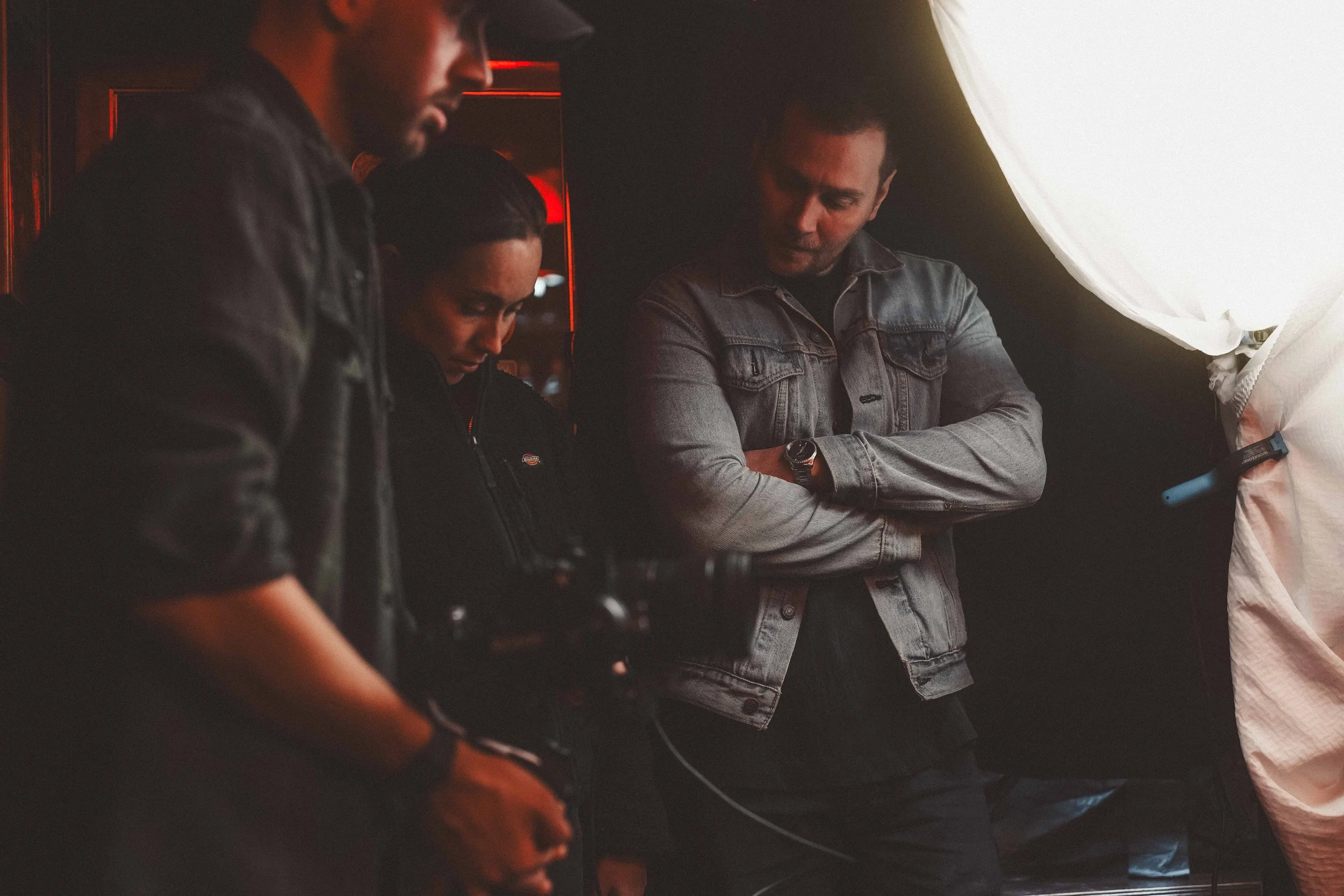 Four people standing close together in a dark room with red and white lights, engaged in a discussion or activity, with one person holding a phone.