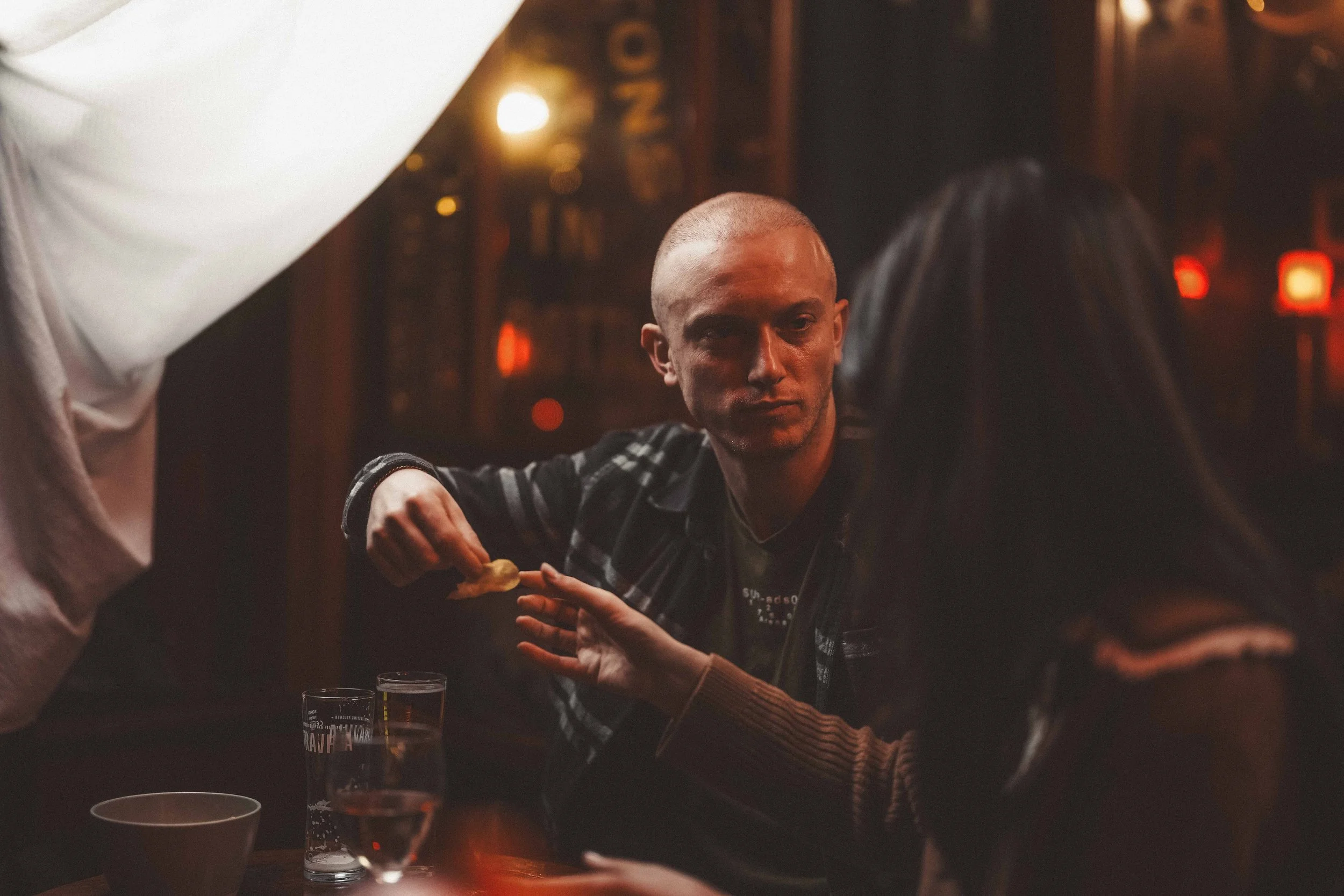 A man with a shaved head and plaid shirt sitting at a dimly lit table in a cozy, warmly decorated restaurant or bar, engaged in conversation with a woman. Glasses and cups are on the table, with a background of warm light and decor.