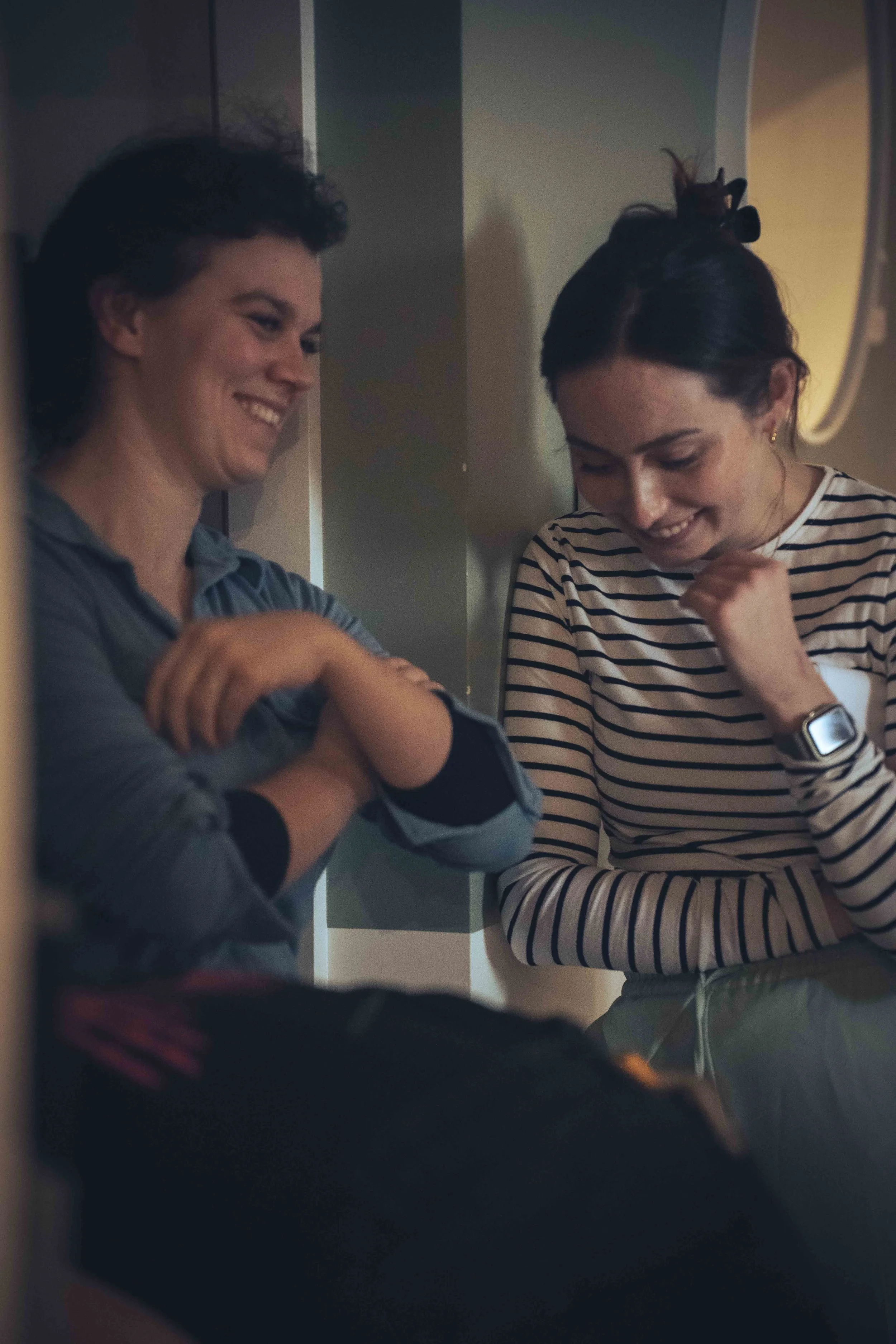 Two women are standing close together, smiling and laughing. One woman has dark, curly hair and is wearing a striped shirt, with a bow in her hair. The other woman has short dark hair and is also smiling, wearing a shirt with a collar. They appear to