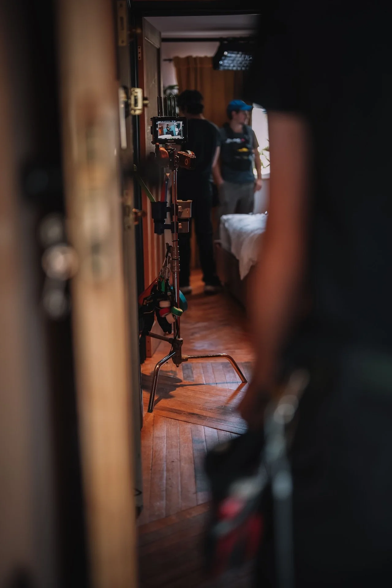 A film or video production scene viewed through a narrow opening, showing two people standing in a room with wooden floors and a bed. One person wears a blue cap, and the other has dark, curly hair. A camera on a tripod is set up in the foreground.