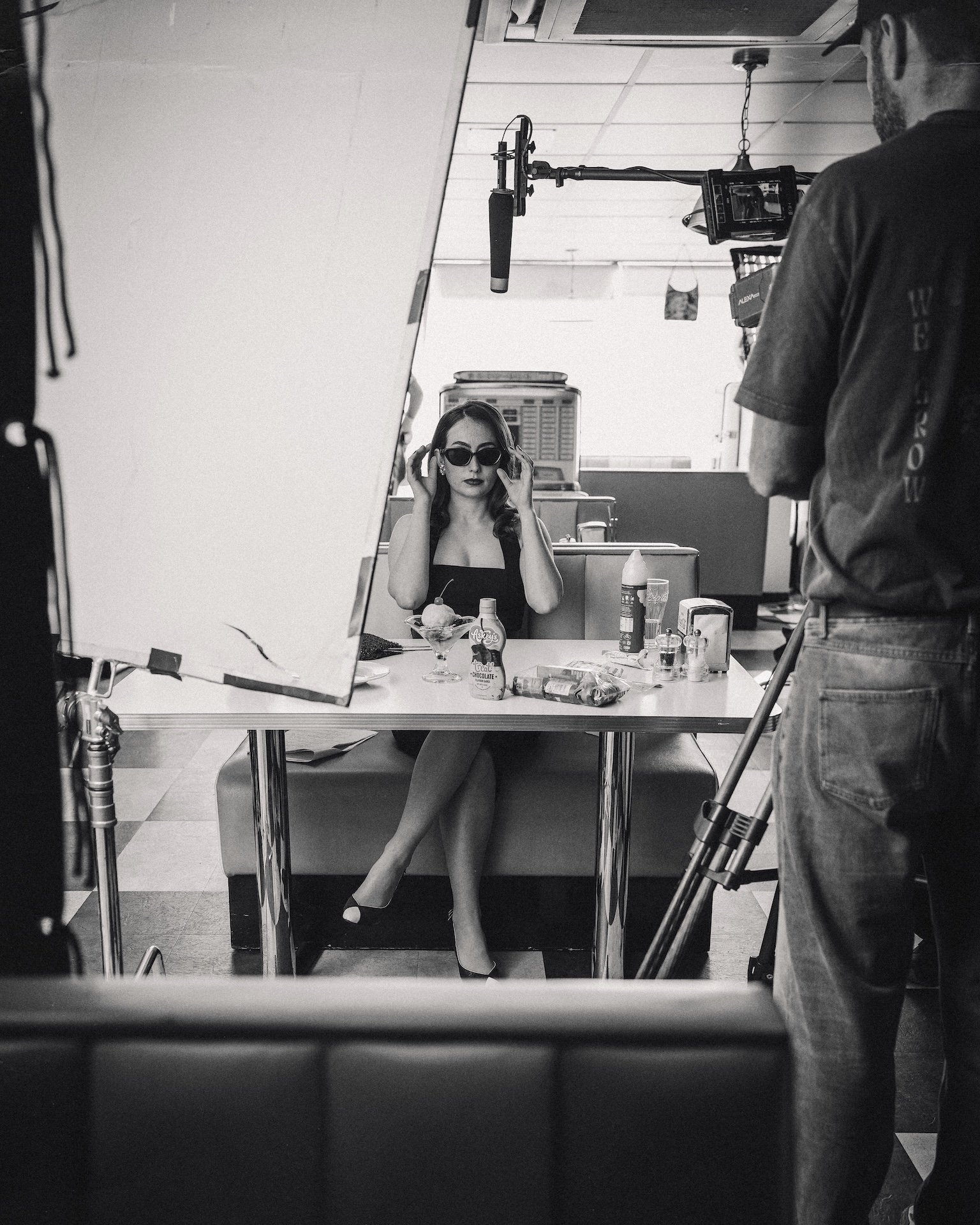 A woman in vintage style with sunglasses, sitting at a table in a diner, with food and condiments in front of her, while a photographer is taking her picture.