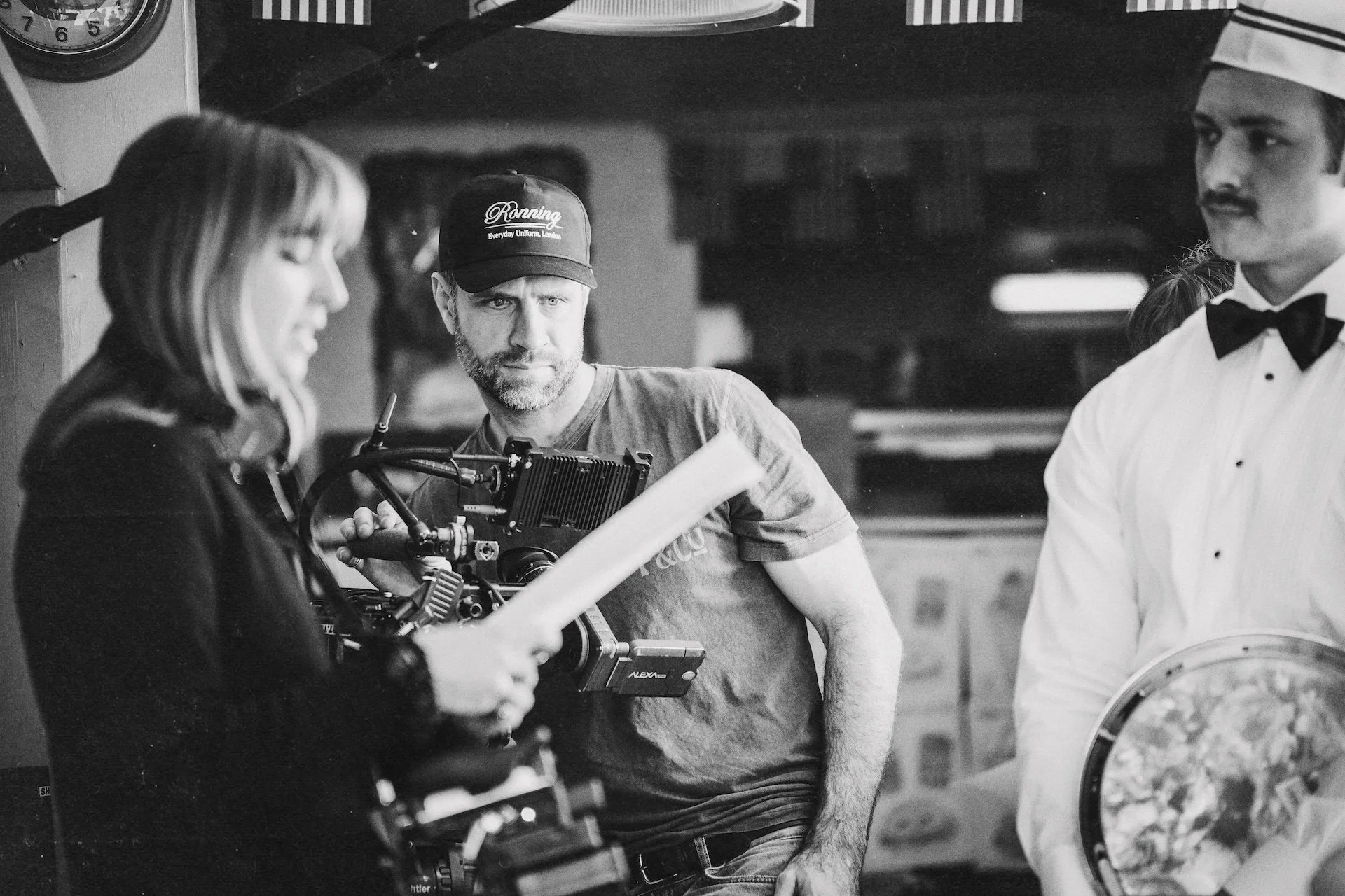A film crew on set, including a woman reviewing notes, a man operating a camera, and another man dressed as a waiter with a bow tie, in a black-and-white shot.