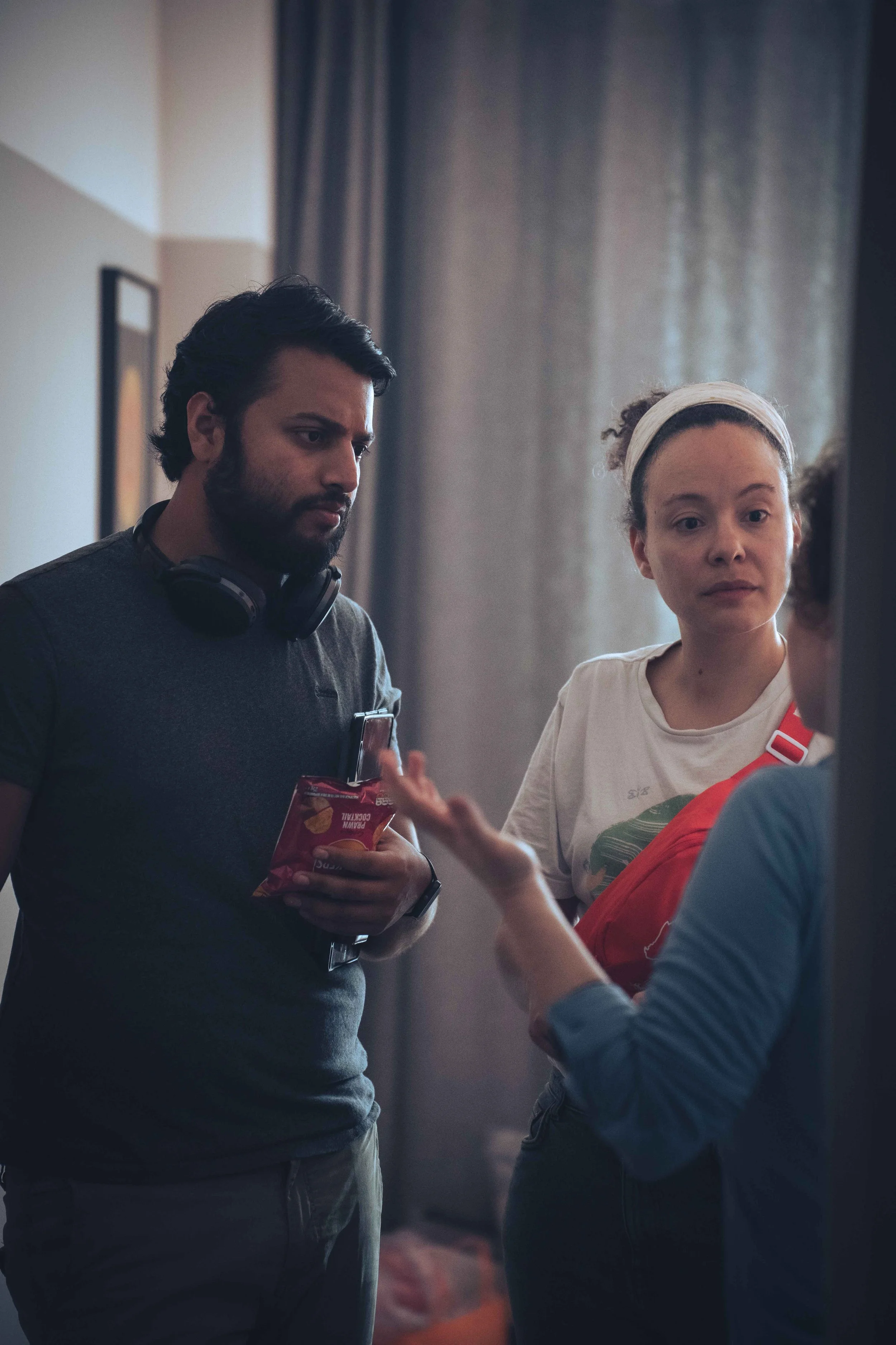 A man with dark hair and a beard, wearing a gray shirt and headphones around his neck, is holding a book or package and looking down. A woman with curly hair, a headband, and a light-colored shirt is next to him, looking forward. They are indoors wit