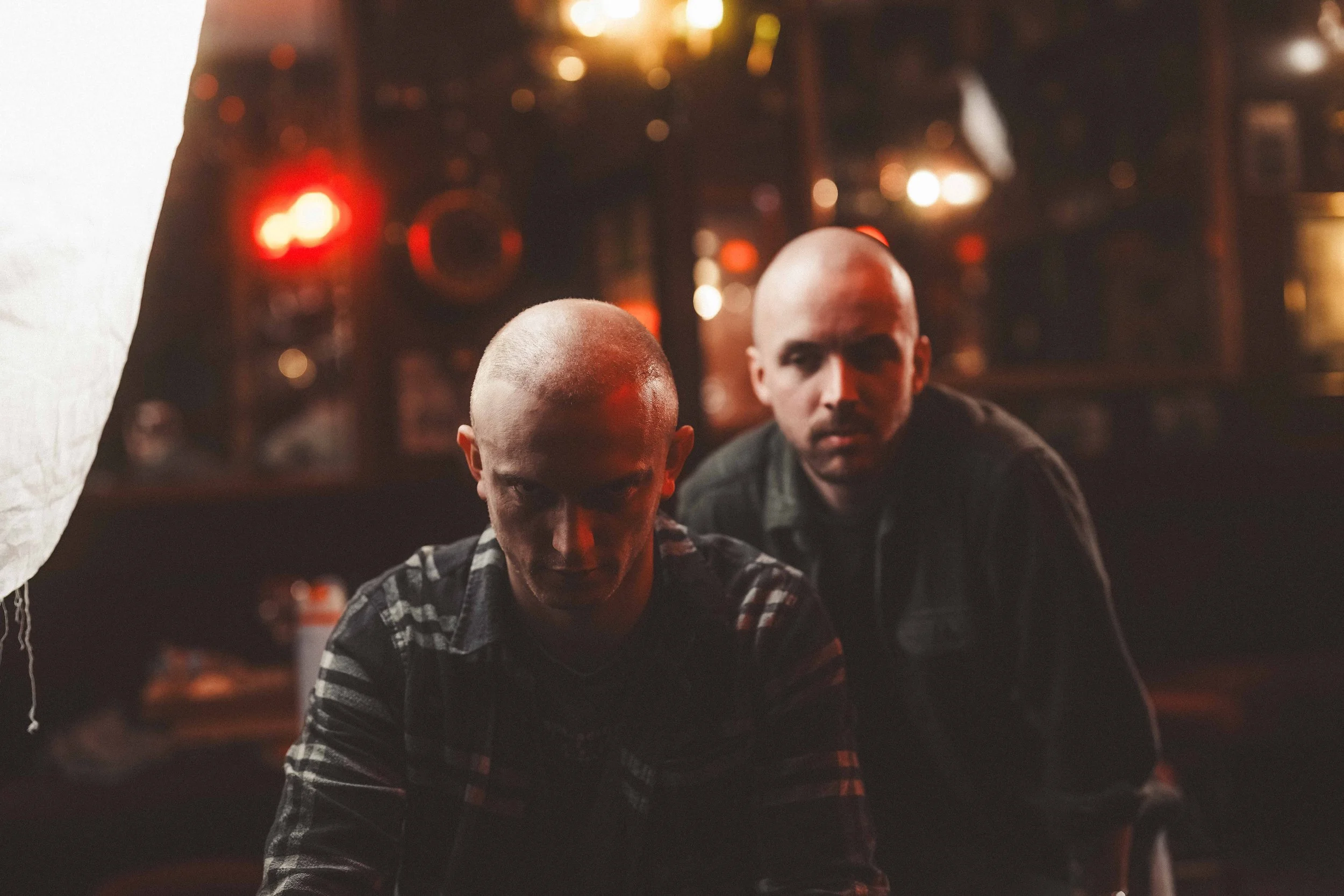 Two bald men sitting in a dimly lit bar or club, with blurred red and yellow lights in the background. One man is wearing a plaid shirt, and the other is looking intently at something.