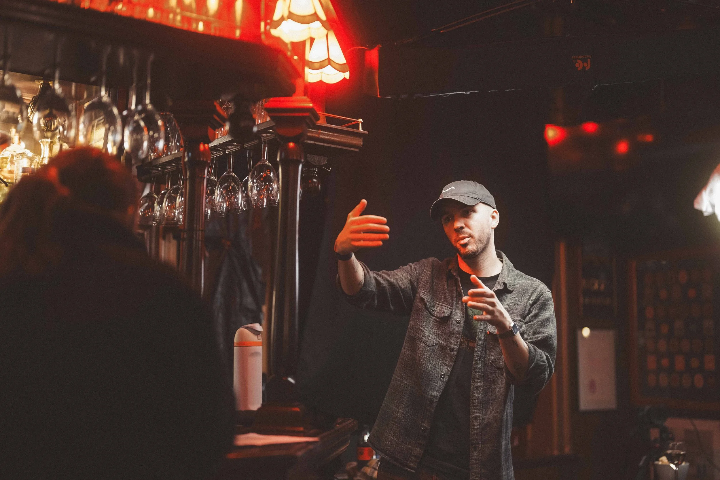 A young man with a beard and wearing a cap is gesturing or explaining something behind a row of hanging wine glasses in a dimly lit bar with red ceiling lights.