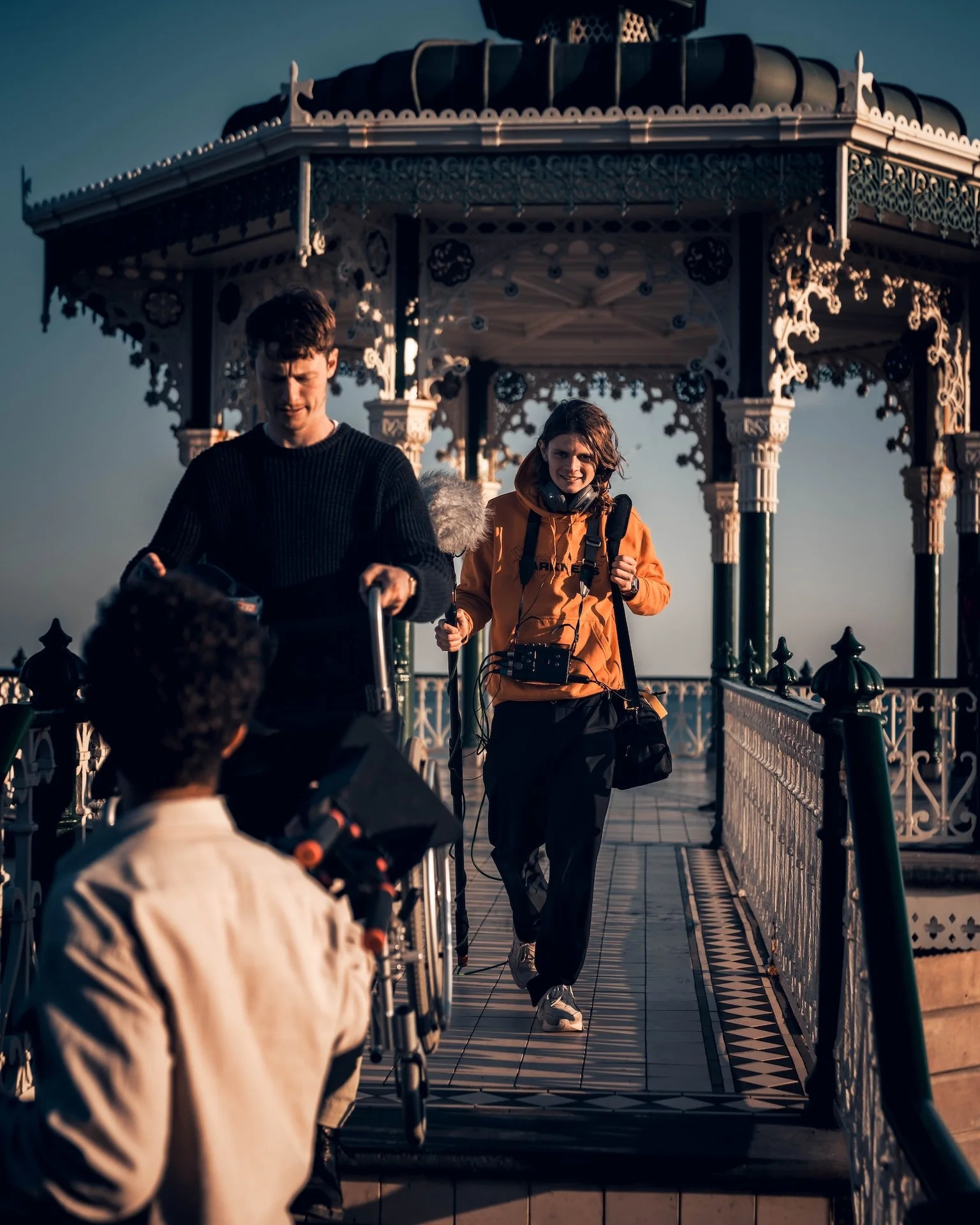 People walking on a decorative bridge with an ornate gazebo in the background during sunset.