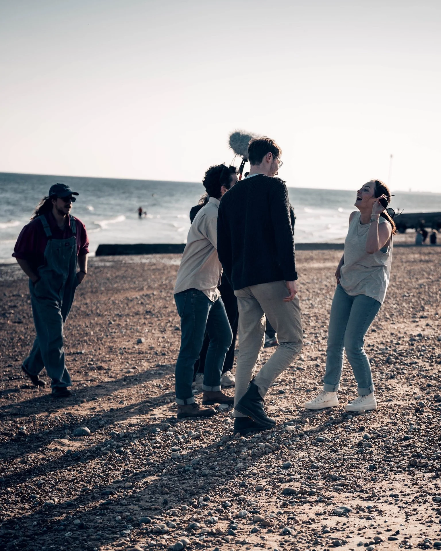 A group of five young adults, four men and one woman, walking and laughing on a pebble beach near the ocean during the daytime. They are casually dressed and appear to be having a fun, relaxed time.