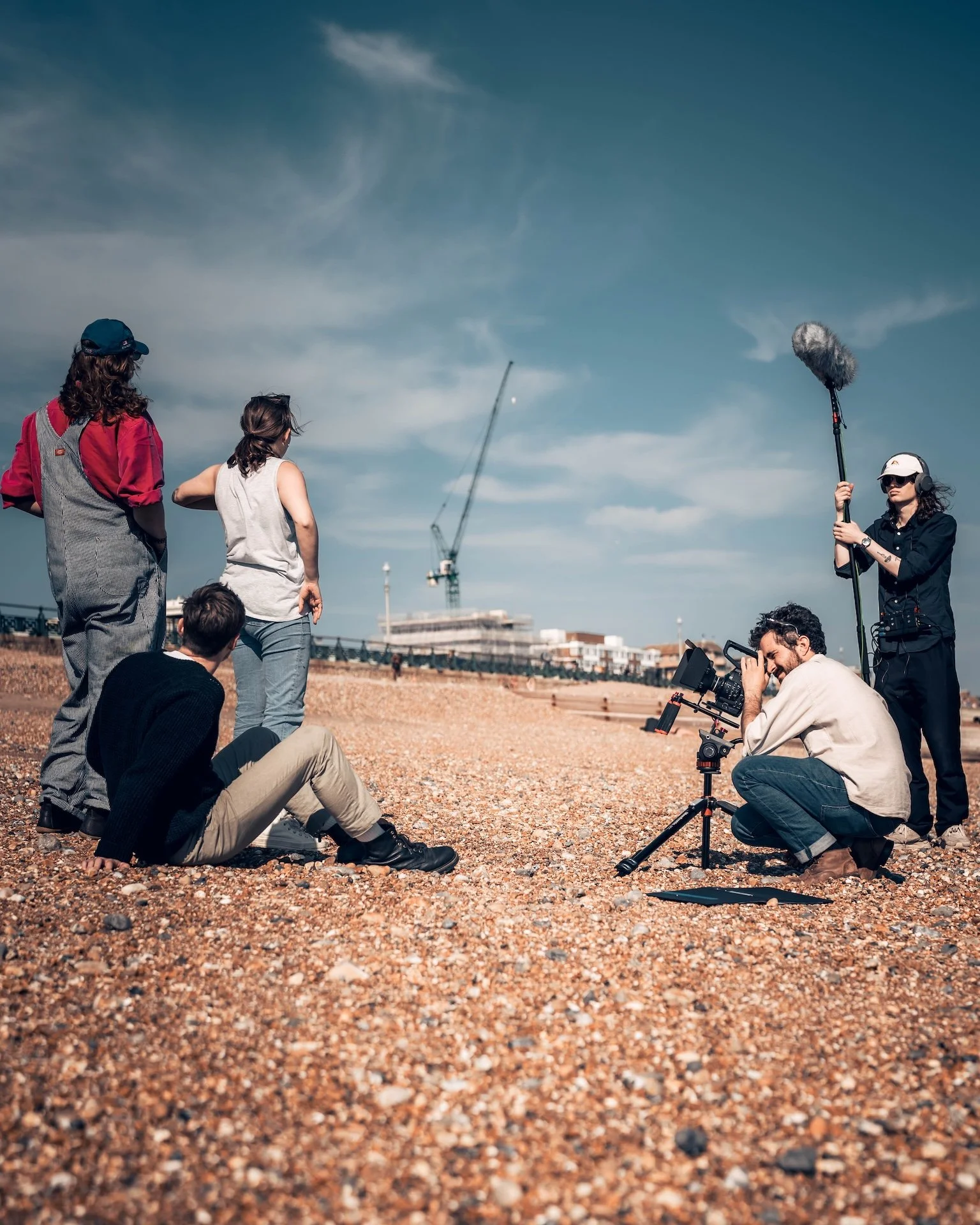 A film crew is filming on a gravel outdoor location with a crane and camera equipment. Several crew members are working, including a person with a boom microphone and another with a camera, while others observe.