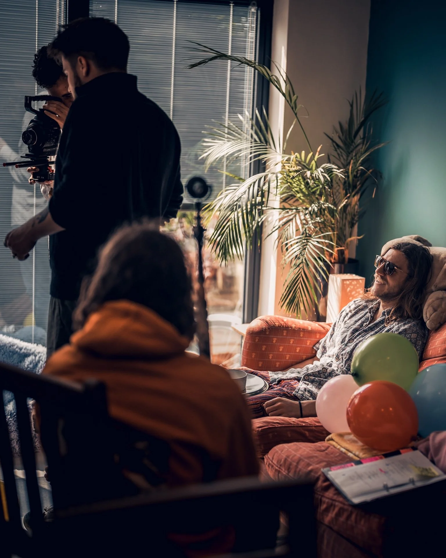 People at a party celebrate around a table with balloons, one man wheres sunglasses and is resting on an orange couch, another person is holding a camera, and a woman with long dark hair is sitting with others in a cozy room with plants and large win