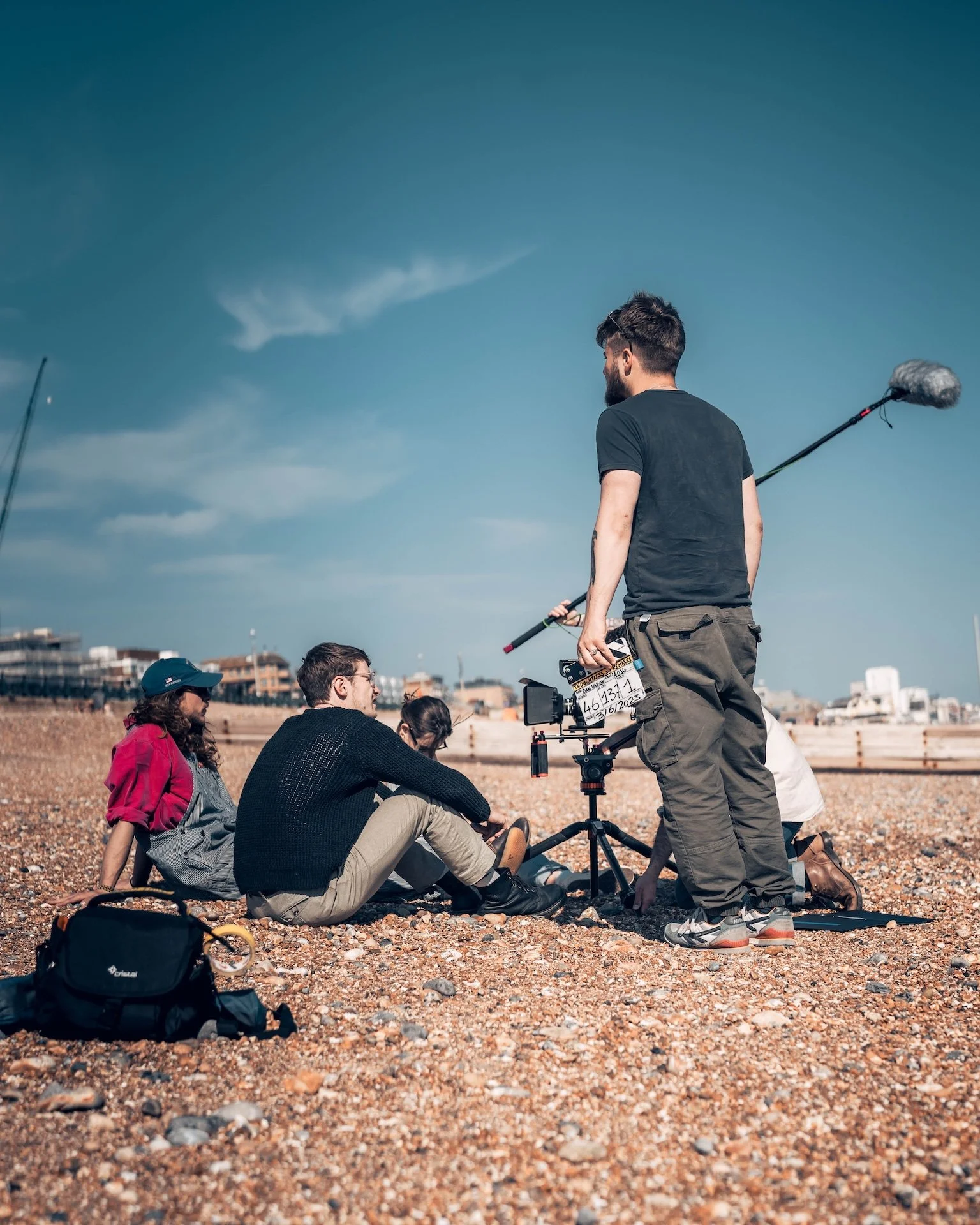 Film crew recording a scene on a pebble beach with a cityscape in the background on a clear day.