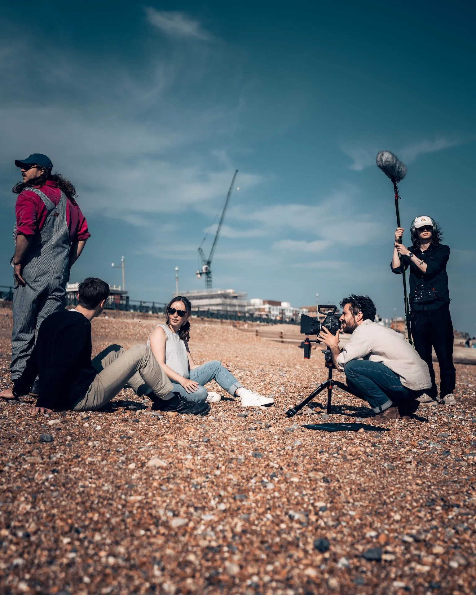 Group of people filming or photographing a woman sitting on a rocky beach, with a person holding a boom microphone and another operating a camera. Construction cranes and buildings are visible in the background under a blue sky.