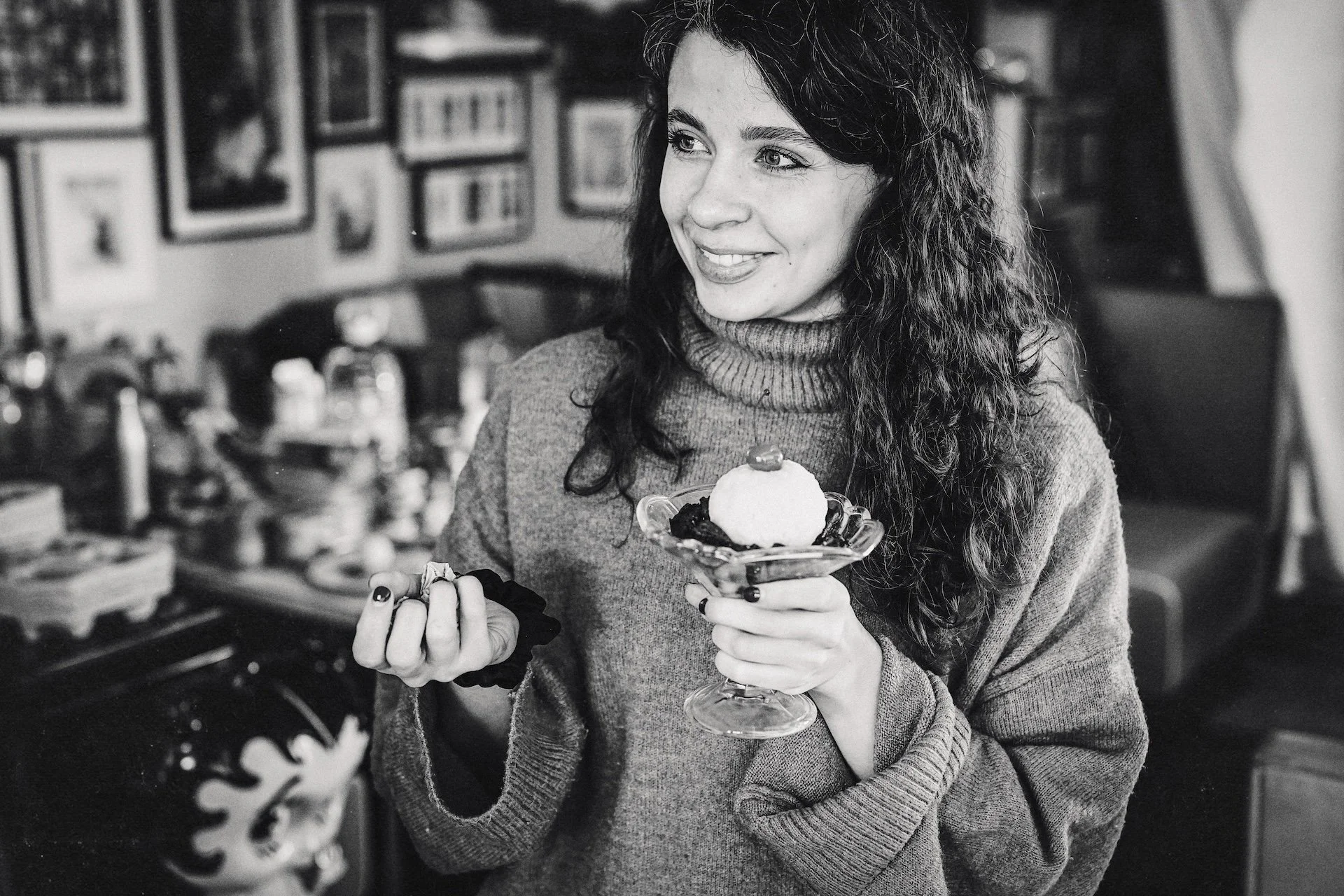 A woman with curly hair holding a dessert glass with ice cream and toppings, smiling in a cozy indoor space with framed pictures on the wall.
