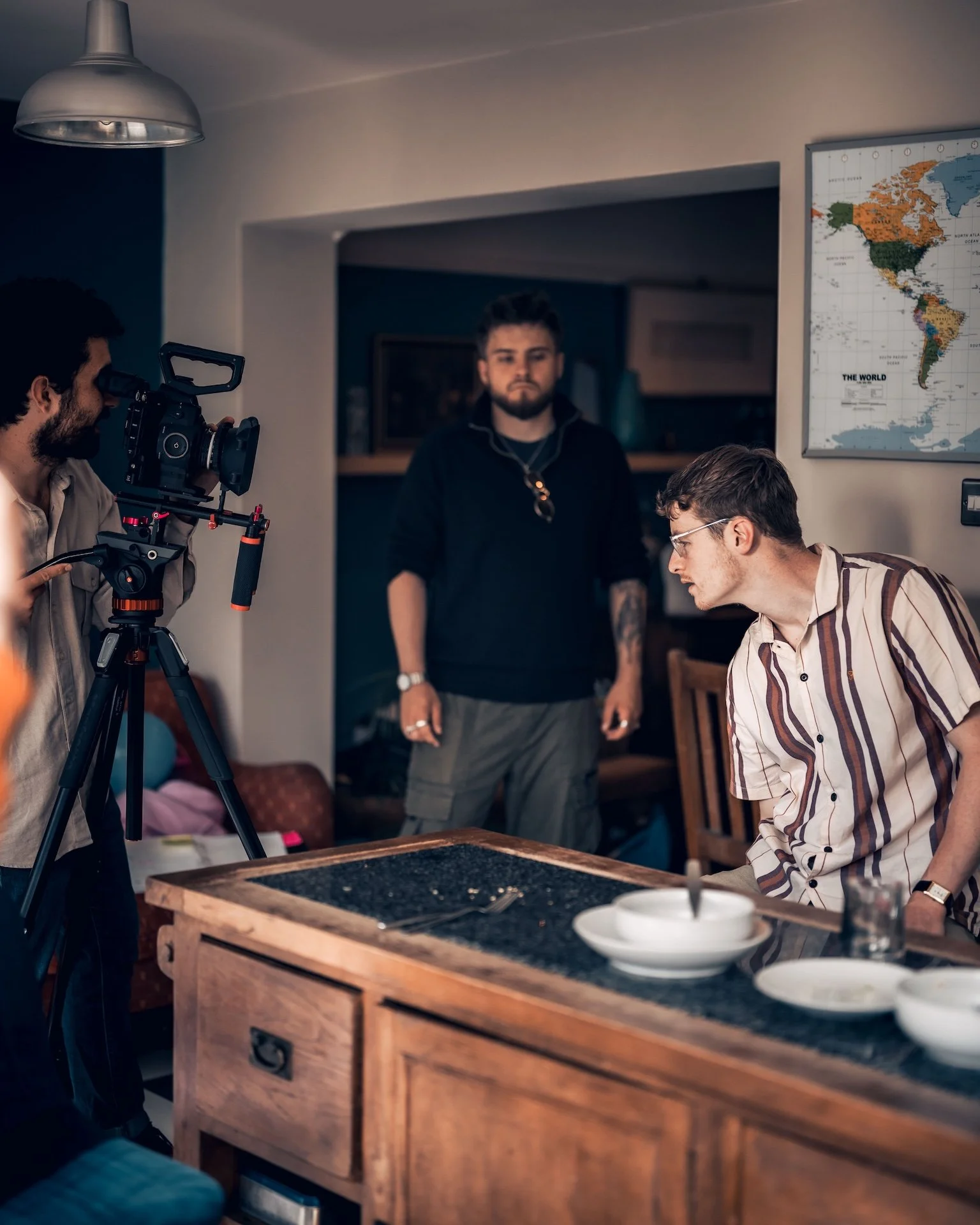 A group of people is filming a scene in a cozy indoor setting with a world map on the wall. One person wearing glasses and a striped shirt is sitting at a table, while another person stands behind him. A camera operator is recording with a profession