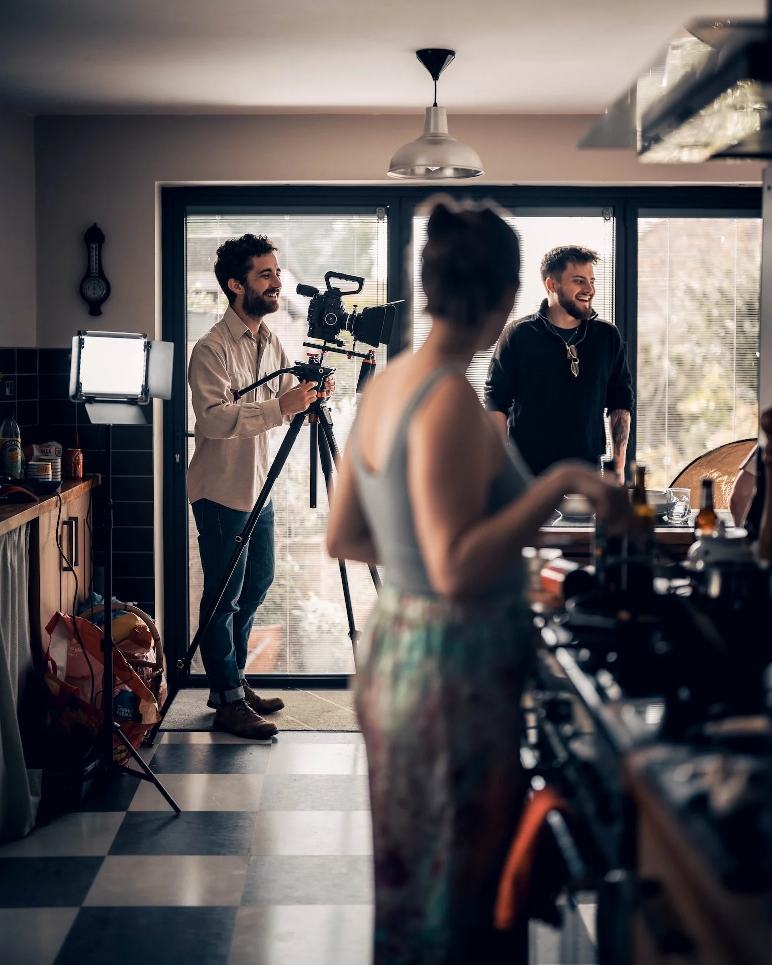 Two smiling men in a kitchen, one with a camera on a tripod and the other wearing a black hoodie, are enjoying a casual conversation. A woman with short hair is partially visible in the foreground.