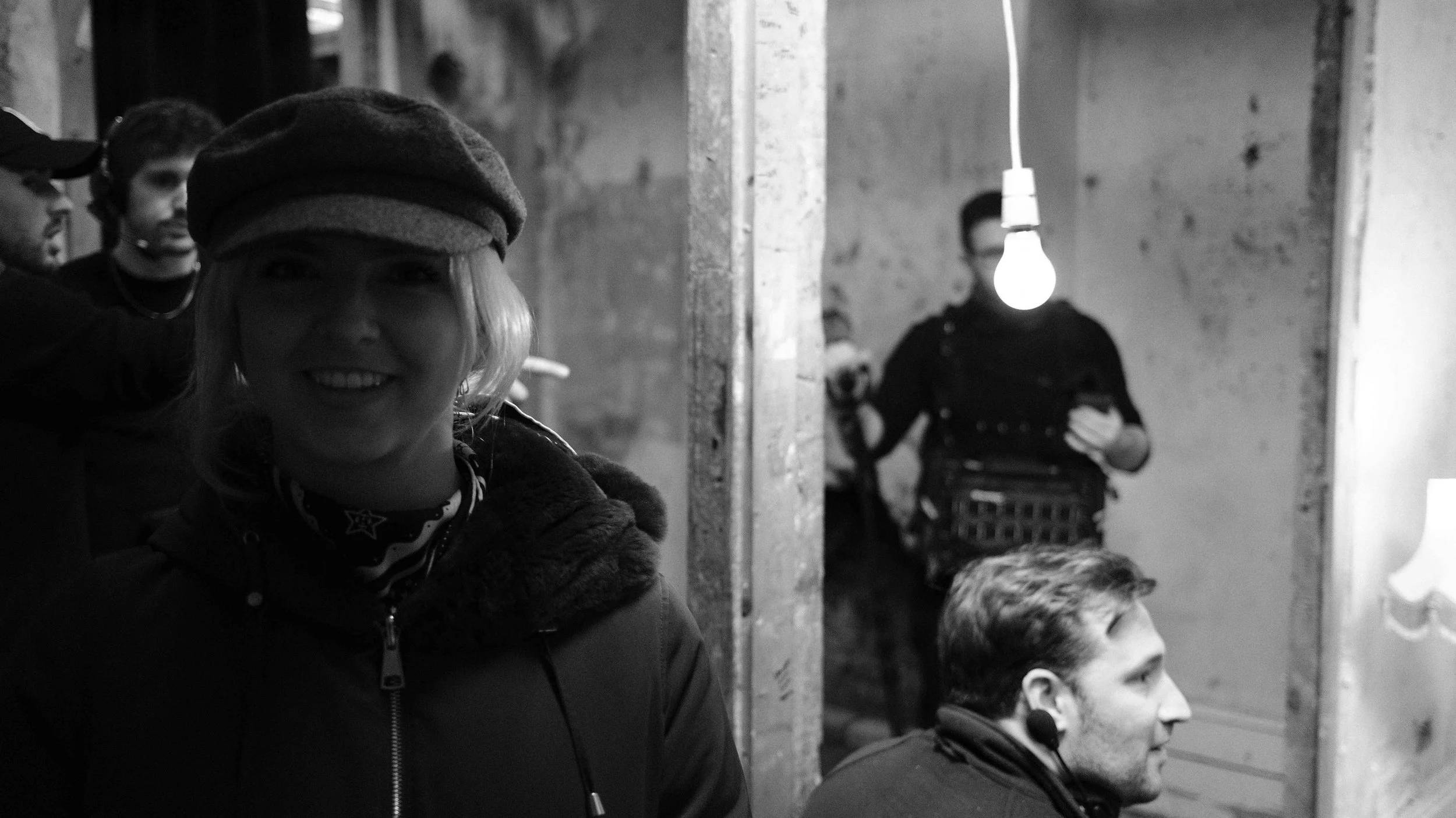 People on a movie set or in a recording studio, with one woman in the foreground smiling, wearing a hat and headphones, and multiple men in the background with headphones and microphones, some holding equipment, in a dimly lit space.
